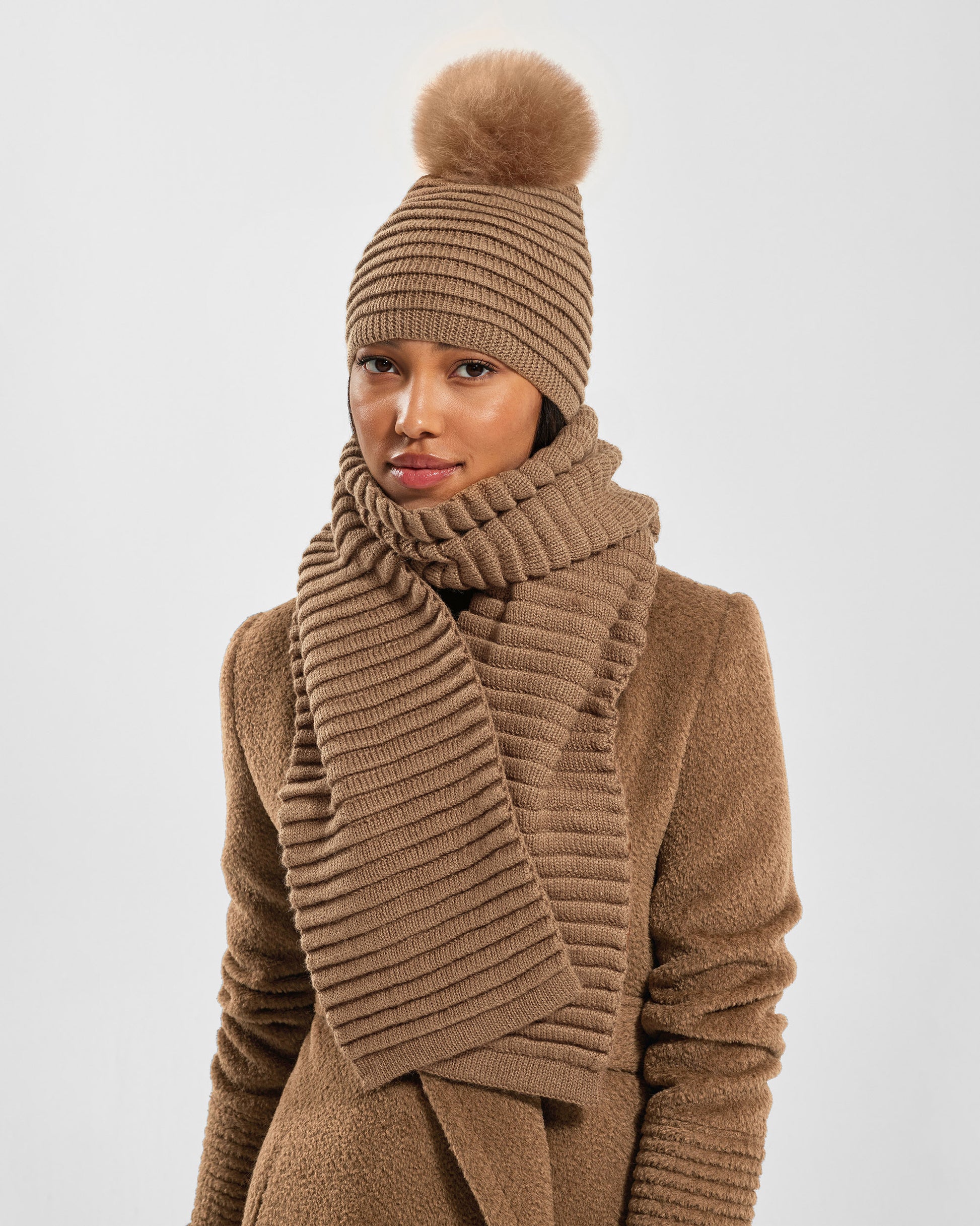 Front view of model wearing Sentaler's adult dark camel ribbed hat with fur pompon, made of baby alpaca yarn paired with a dark camel coat.