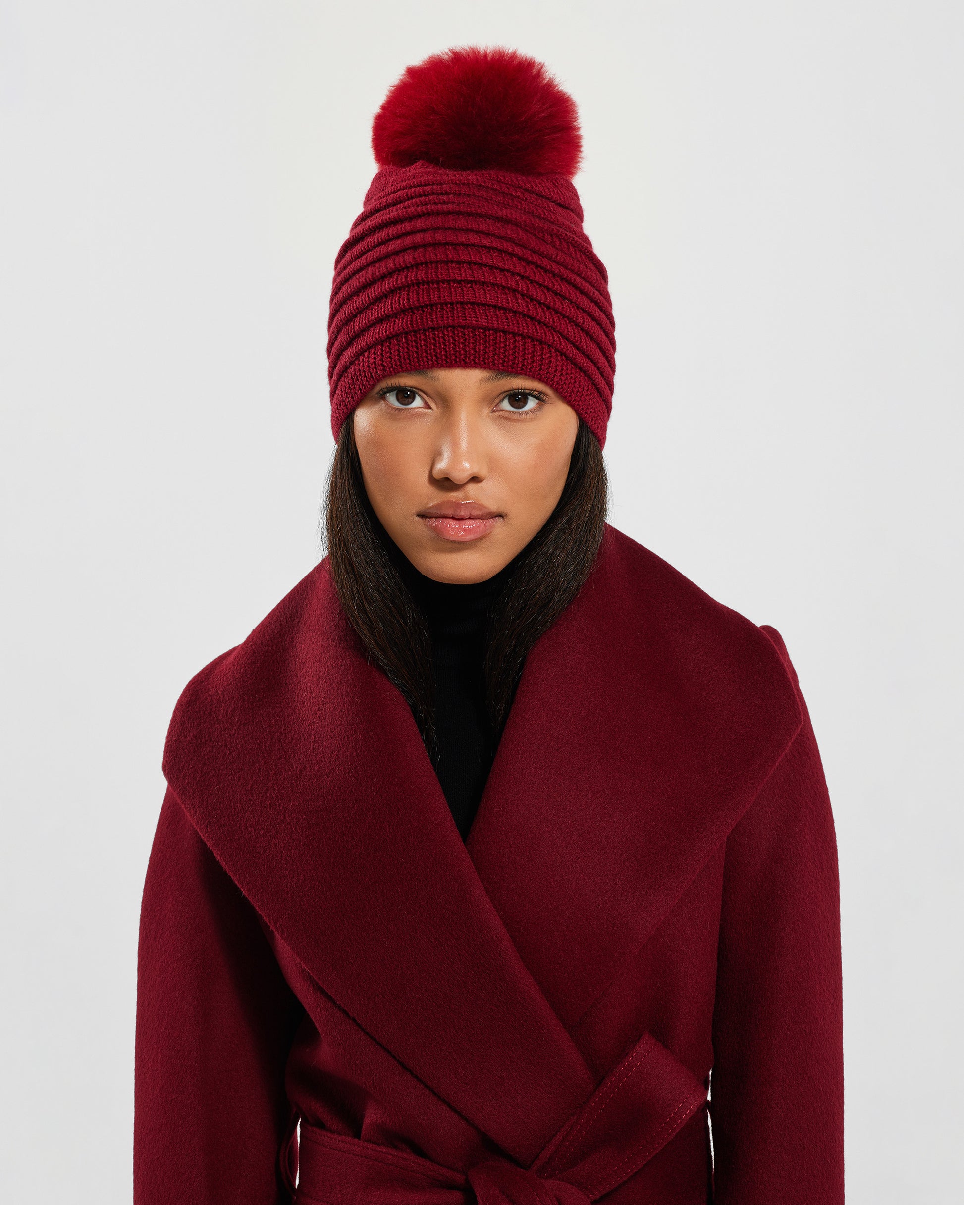 Front view of model wearing Sentaler's adult garnet red ribbed hat with fur pompon, made of baby alpaca yarn, paired with a garnet red coat.