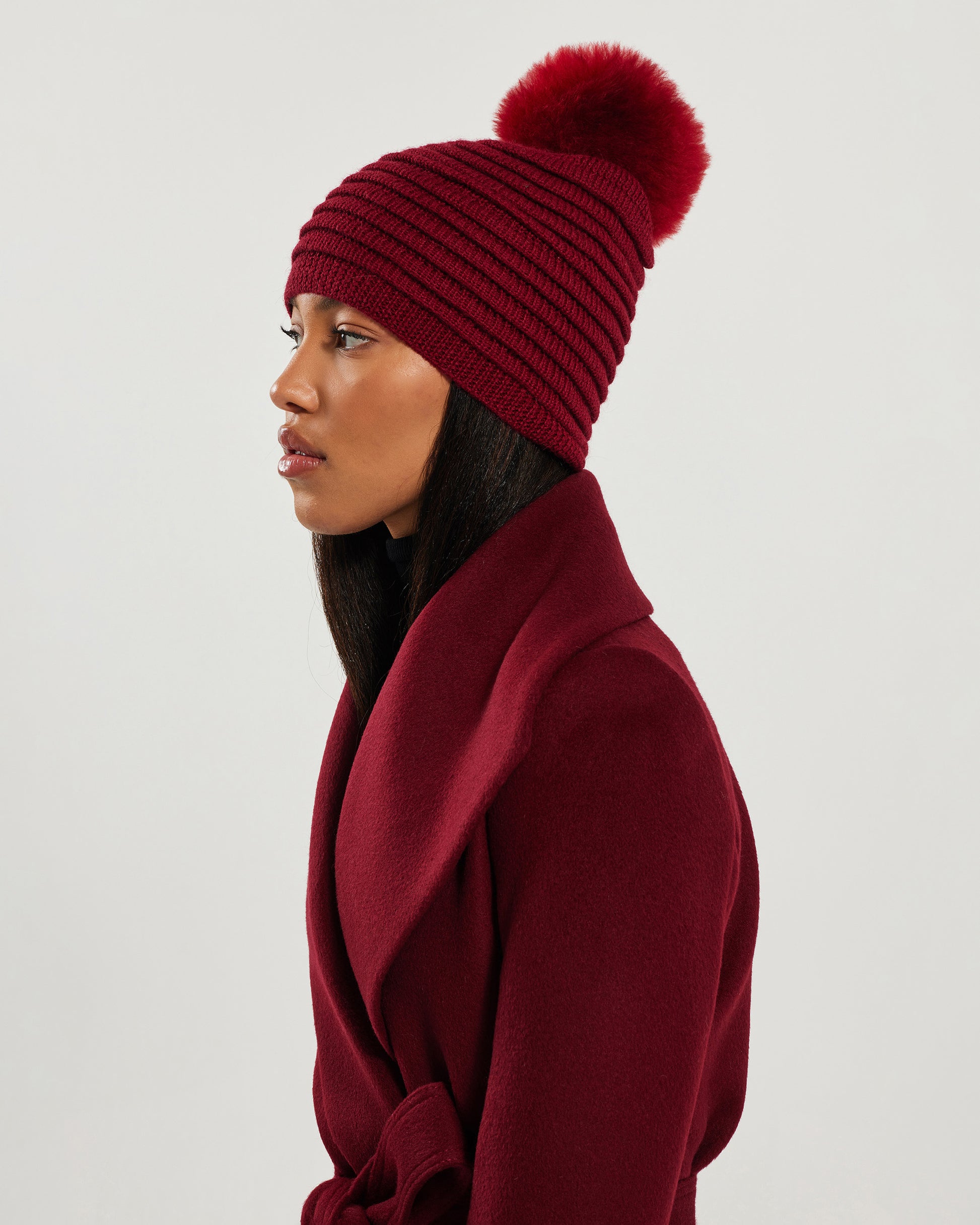 Side view of model wearing Sentaler's adult garnet red ribbed hat with fur pompon, made of baby alpaca yarn, paired with a garnet red coat.