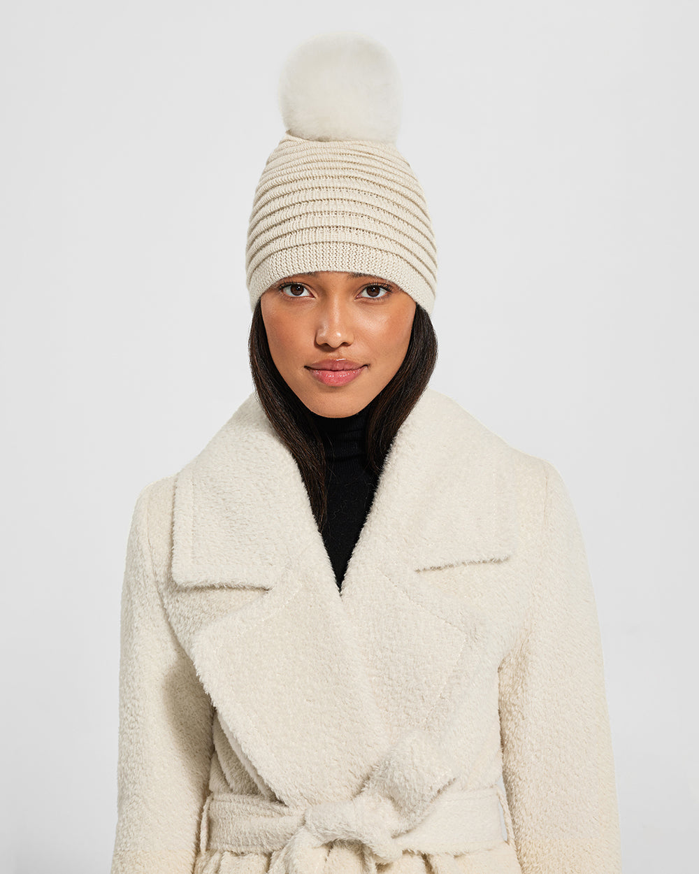 Front view of model wearing Sentaler's adult ivory ribbed hat with fur pompon, made of baby alpaca yarn, paired with an ivory coat.