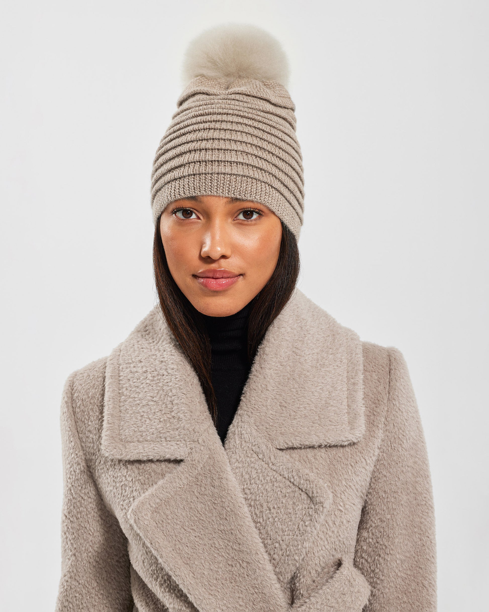 Front view of model wearing Sentaler's adult light taupe ribbed hat with fur pompon, made of baby alpaca yarn, paired with a light taupe coat.
