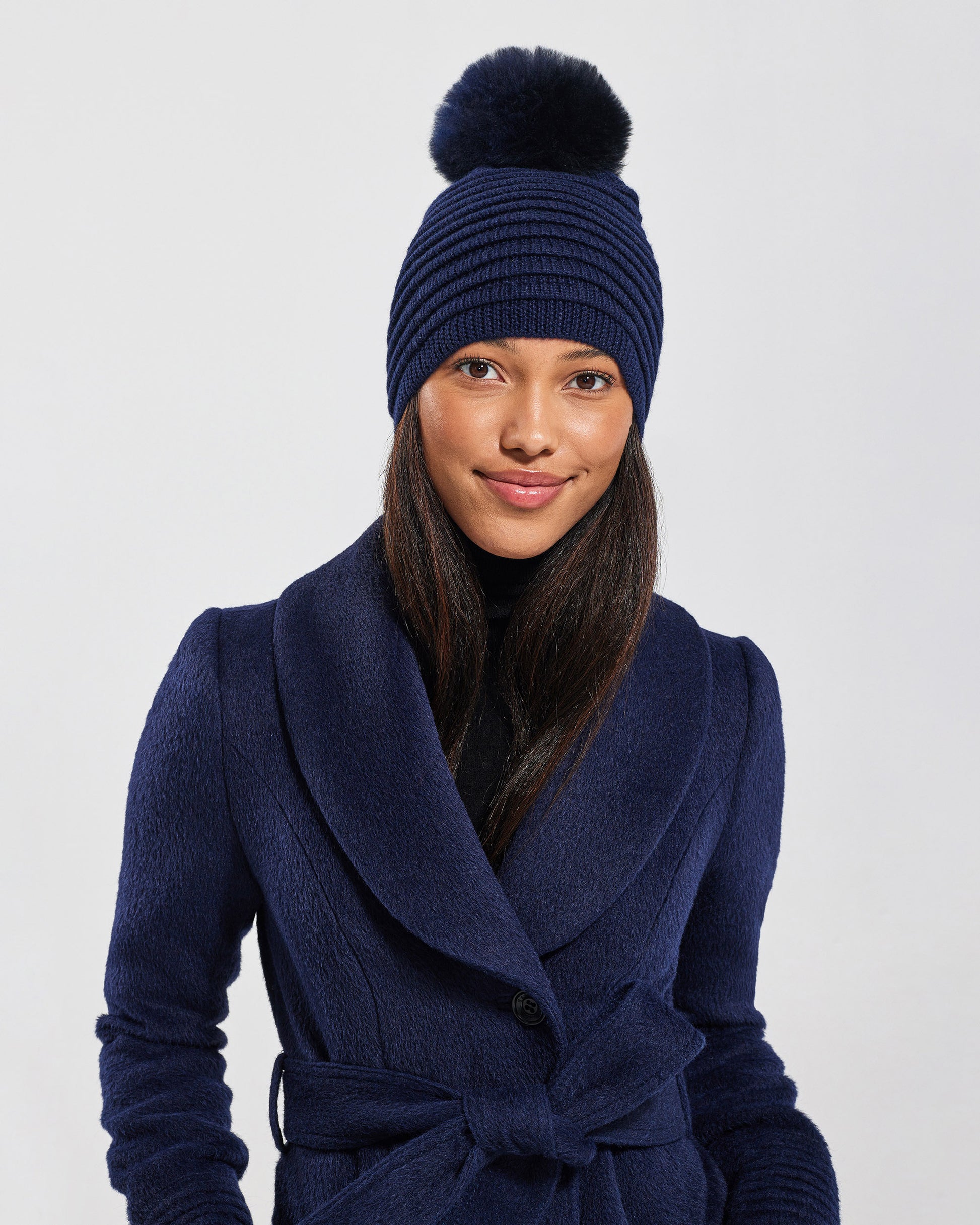 Front view of model wearing Sentaler's adult navy ribbed hat with fur pompon, made of baby alpaca yarn, paired with a deep navy coat.