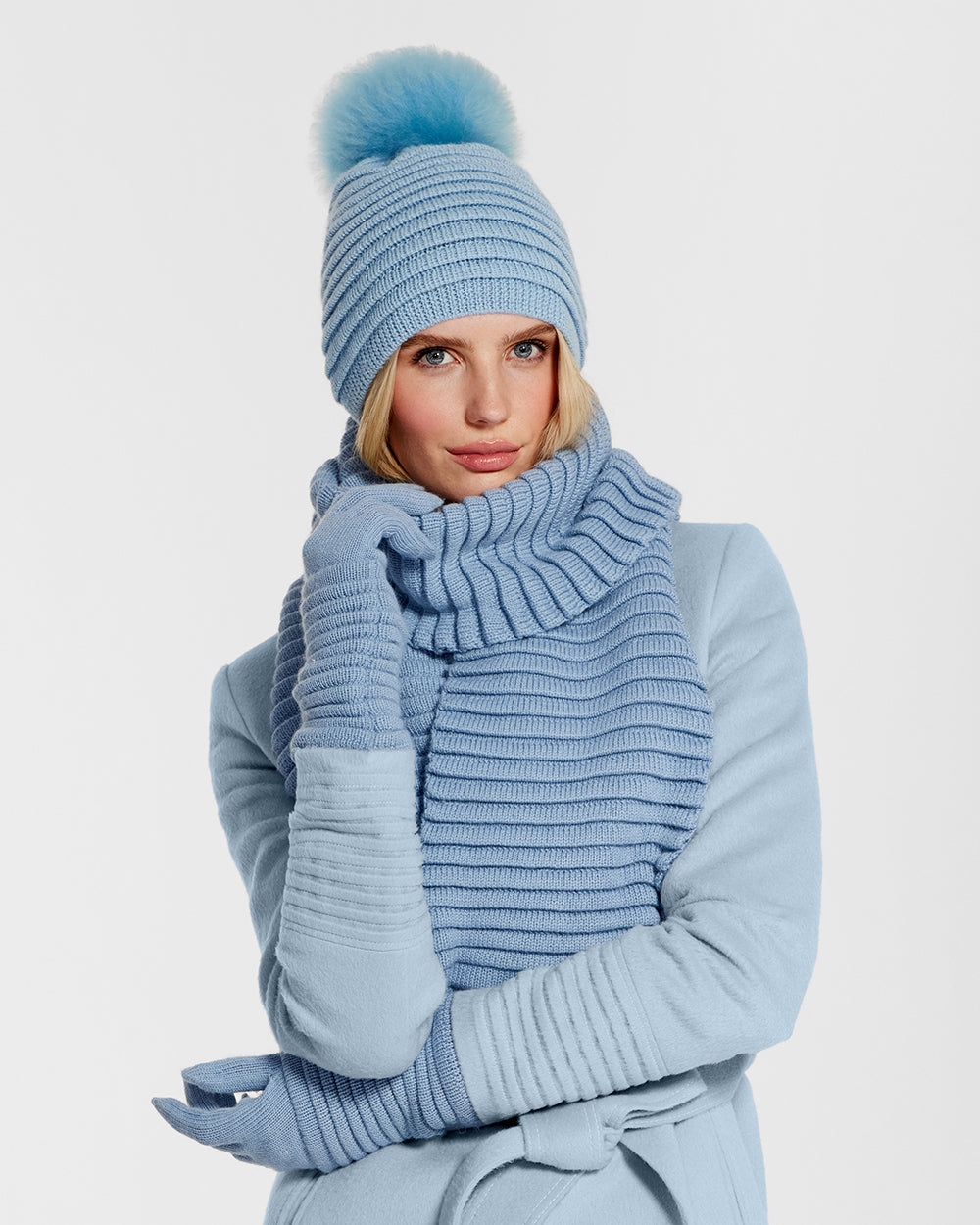 Sentaler Adult Ribbed Powder Blue Hat with Oversized Fur Pompon, Adult Ribbed Powder Blue Scarf and Adult Ribbed Powder Blue Gloves in Baby Alpaca yarn. Seen from front close up on female model.