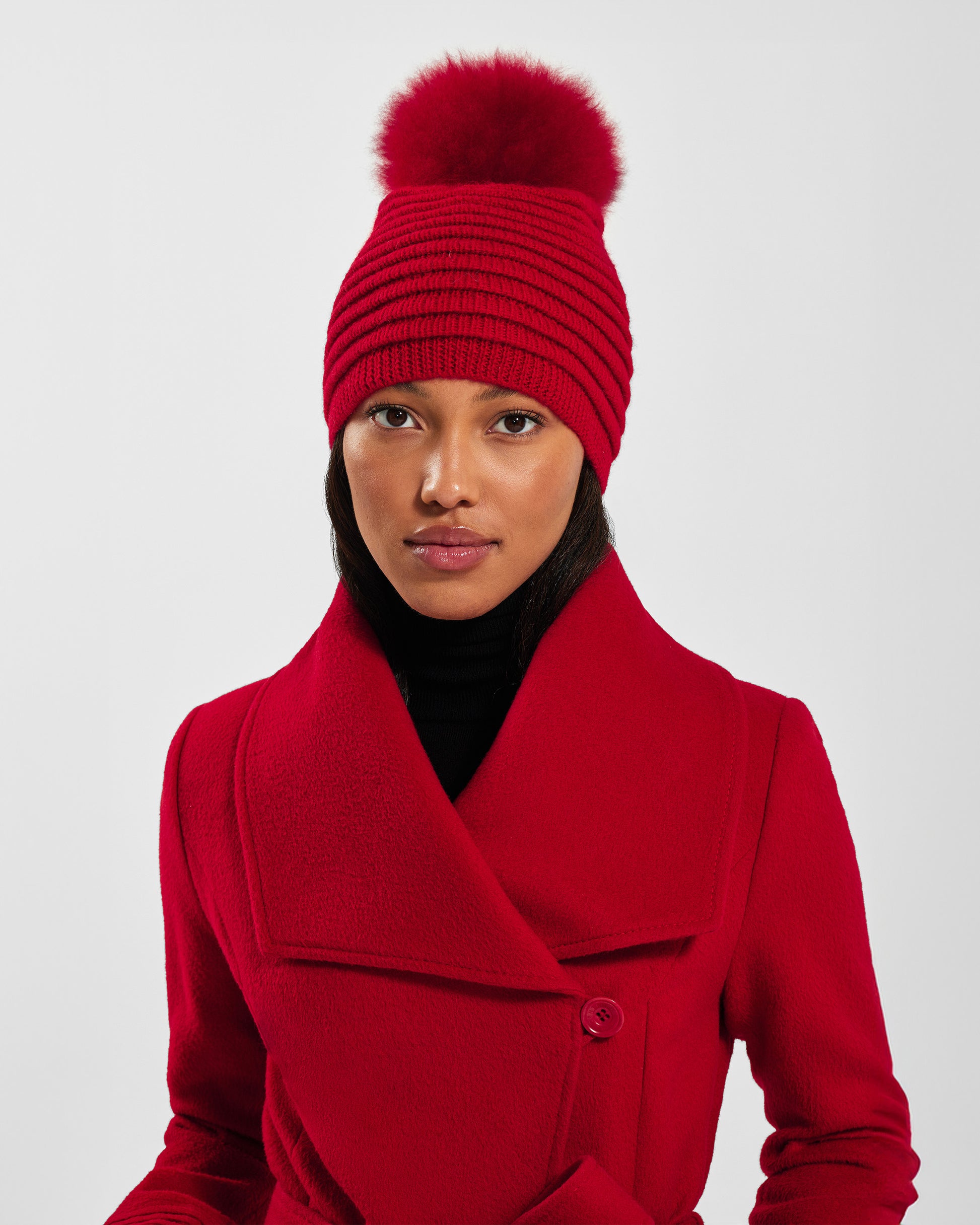 Front view of model wearing Sentaler's adult red ribbed hat with fur pompon, made of baby alpaca yarn, paired with a scarlet red coat.