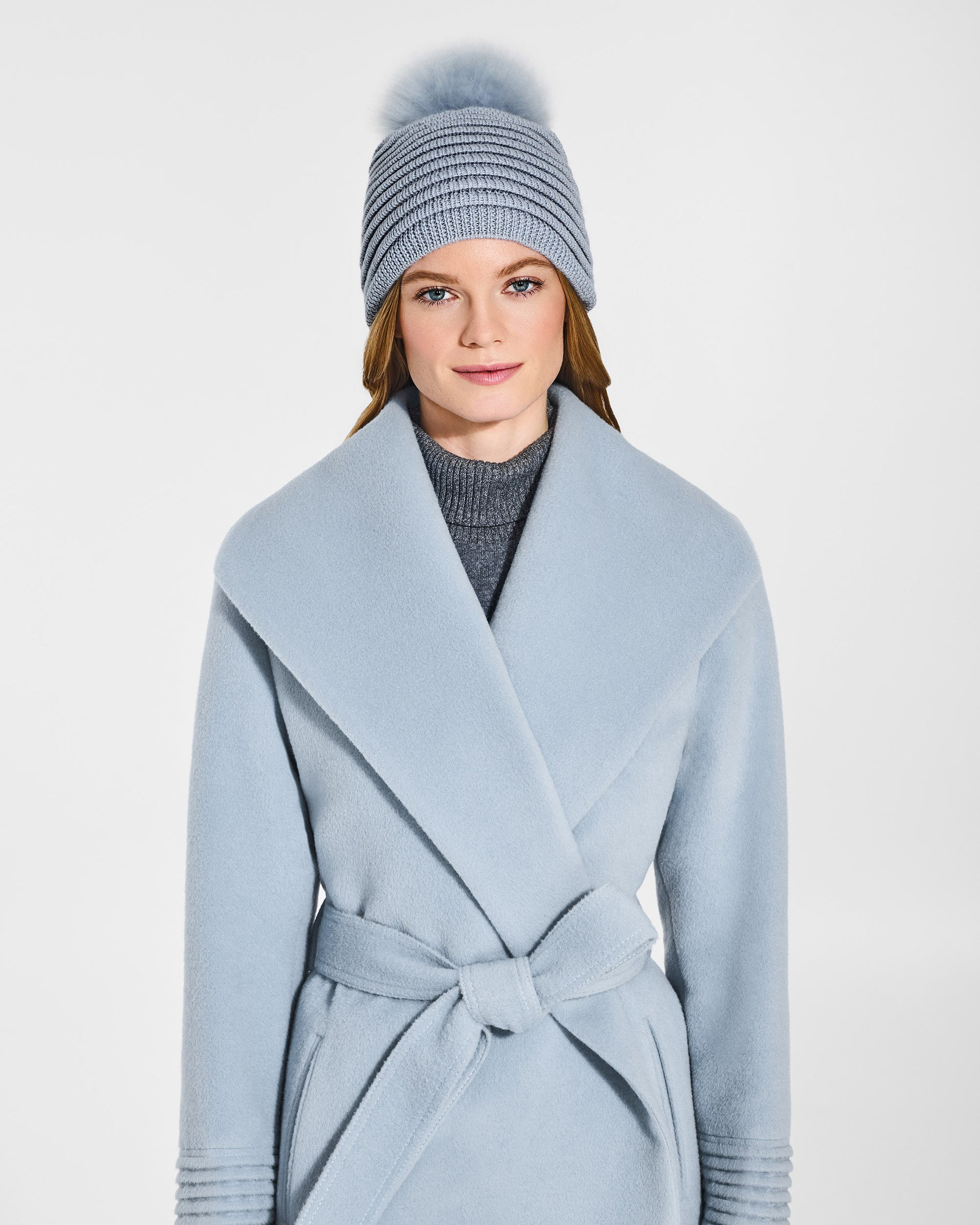 Front view of model wearing Sentaler's adult glacial blue ribbed hat with fur pompon, made of baby alpaca yarn paired with a glacial blue coat.