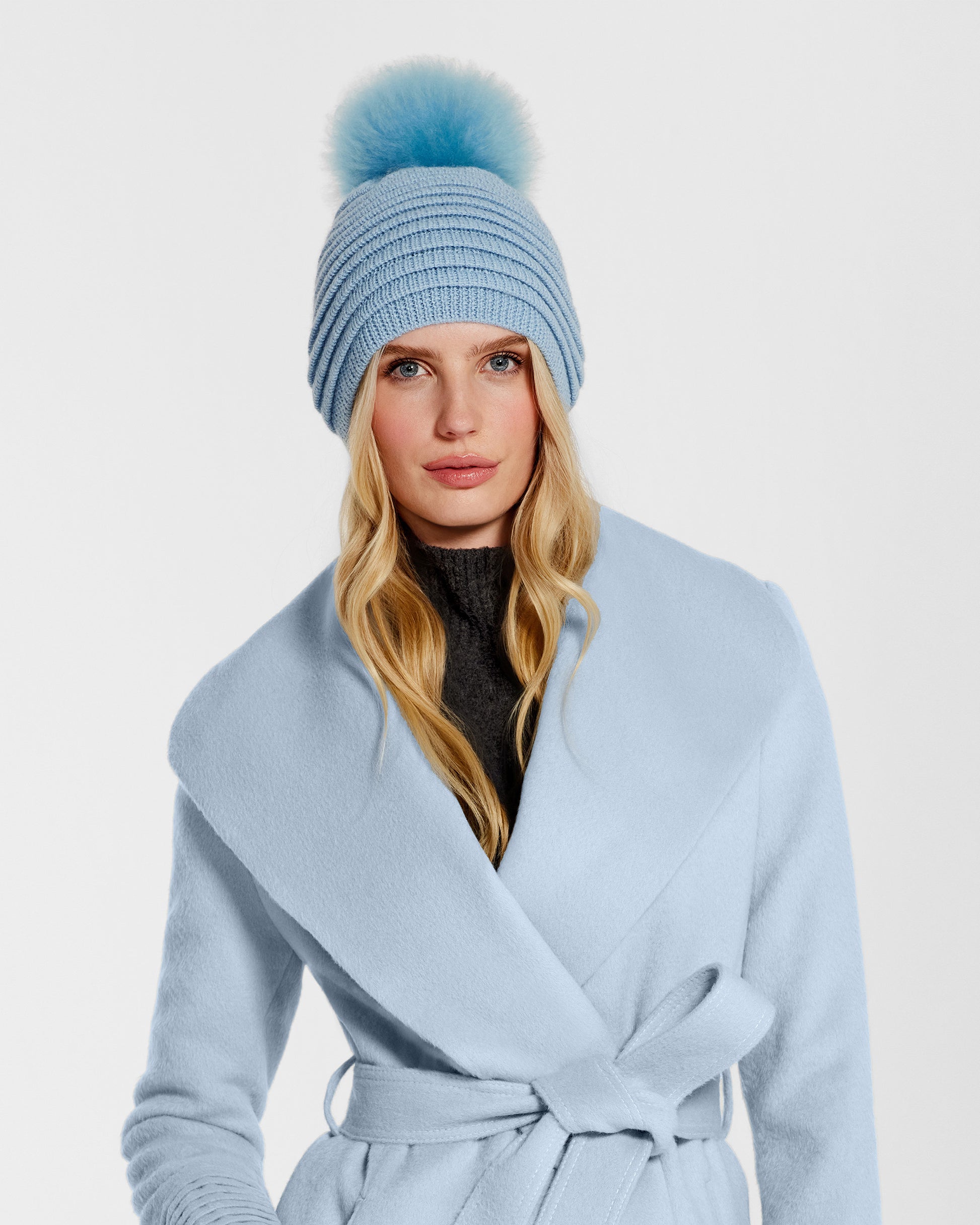 Front view of model wearing Sentaler's adult powder blue ribbed hat with fur pompon, made of baby alpaca yarn, paired with a powder blue coat.