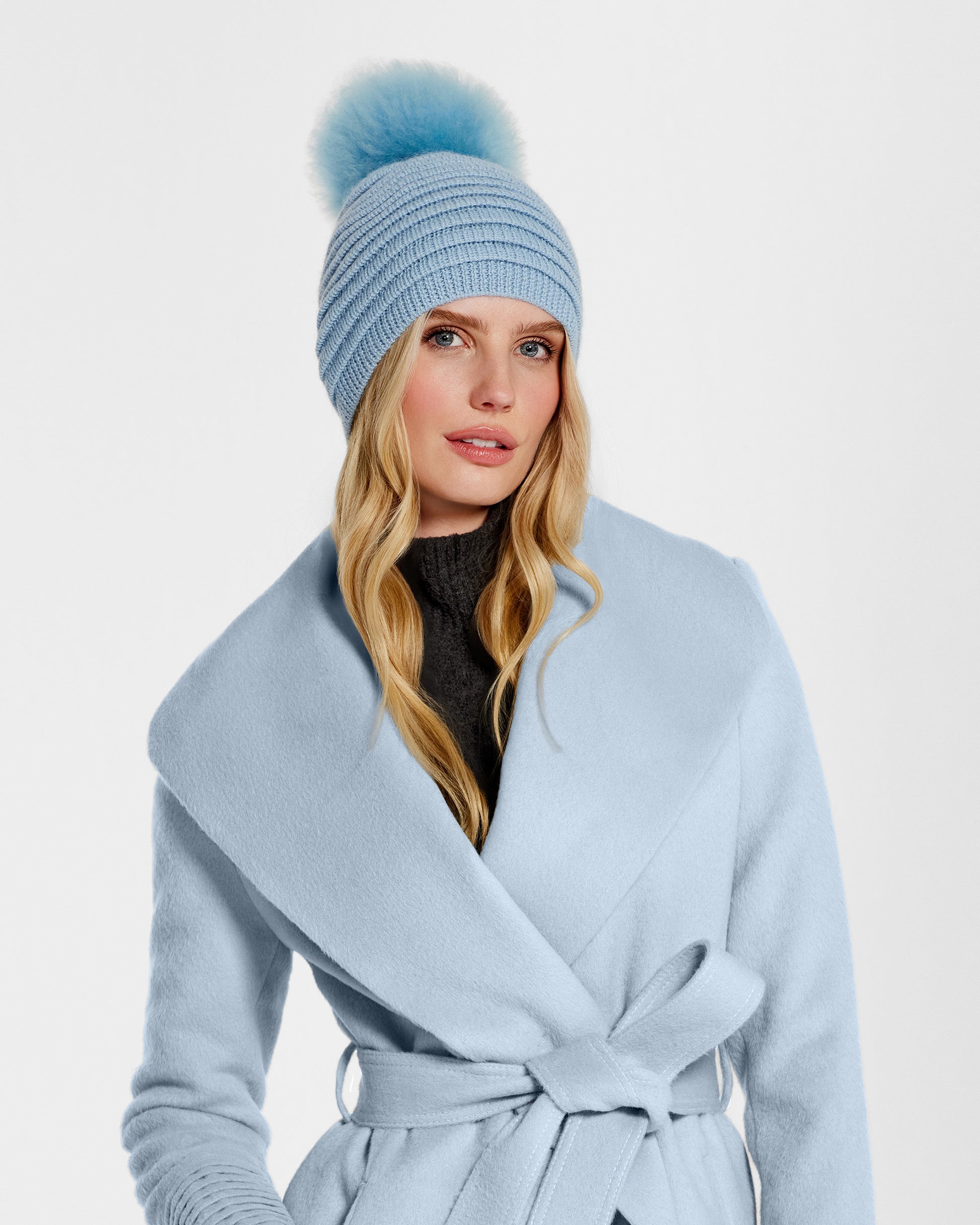 Front view of model wearing Sentaler's adult powder blue ribbed hat with fur pompon, made of baby alpaca yarn, paired with a powder blue coat.