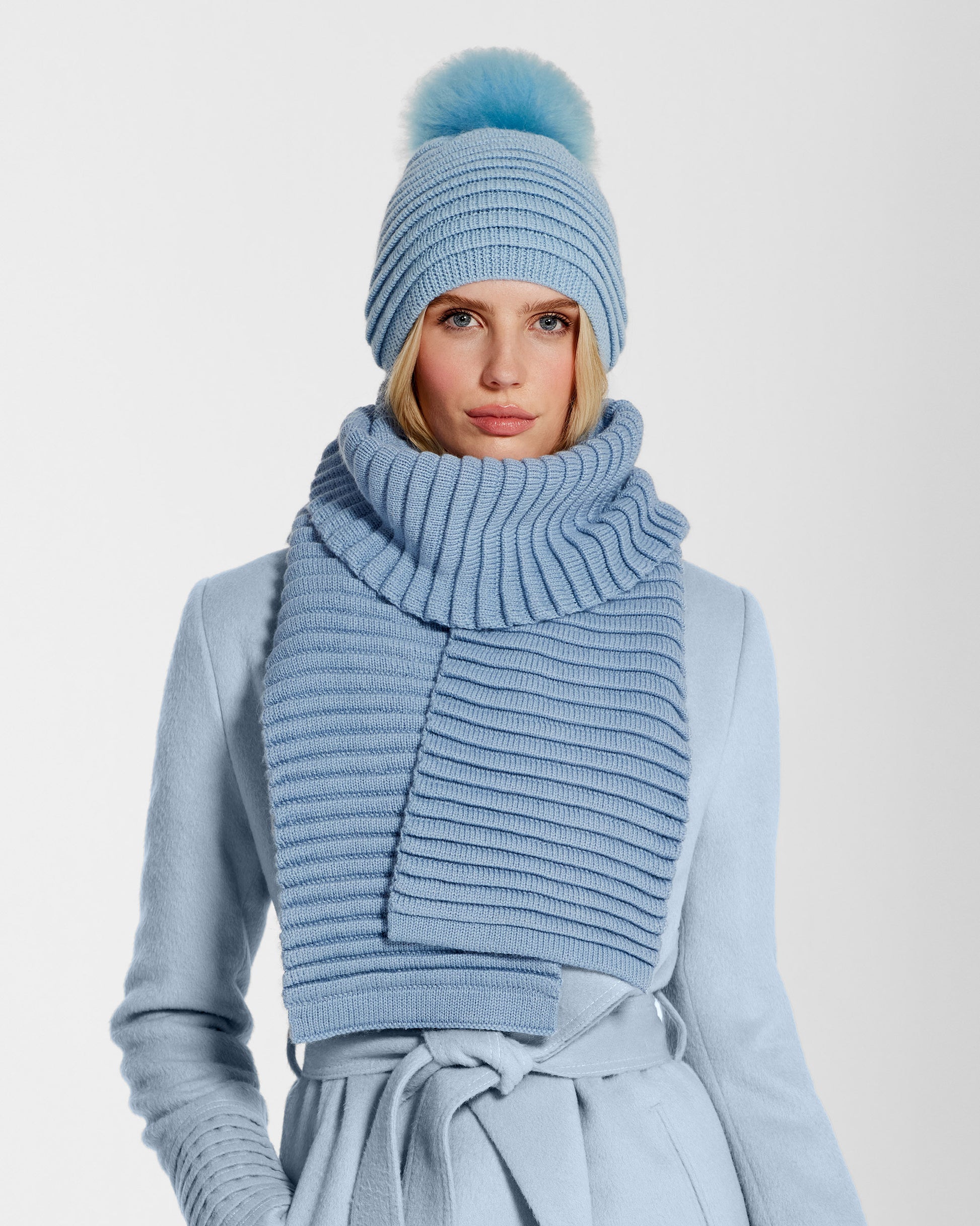 Front view of model wearing Sentaler's adult powder blue ribbed hat with fur pompon, made of baby alpaca yarn, paired with a powder blue coat.