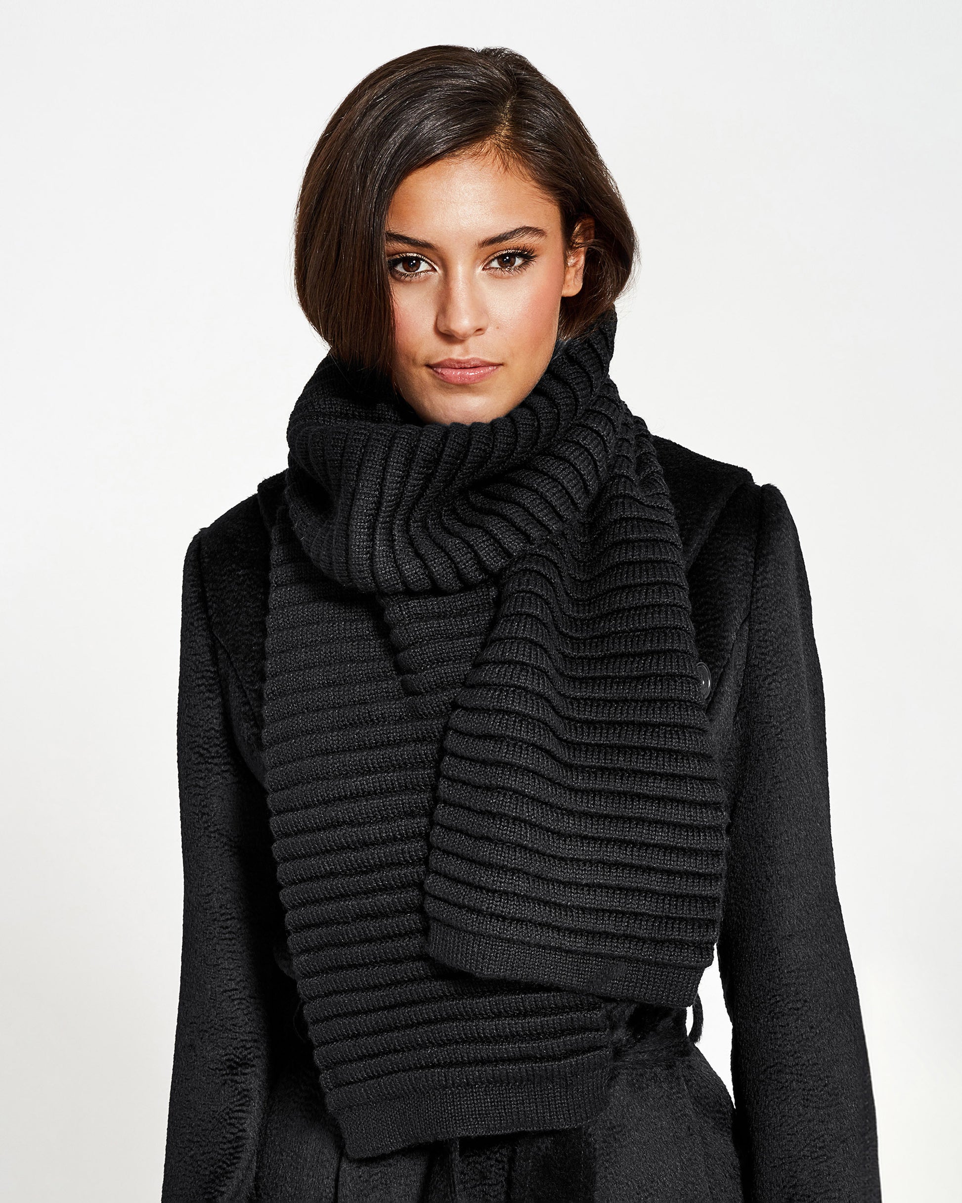 Front view of model wearing Sentaler's adult black ribbed scarf, made of baby alpaca yarn, paired with a black coat.