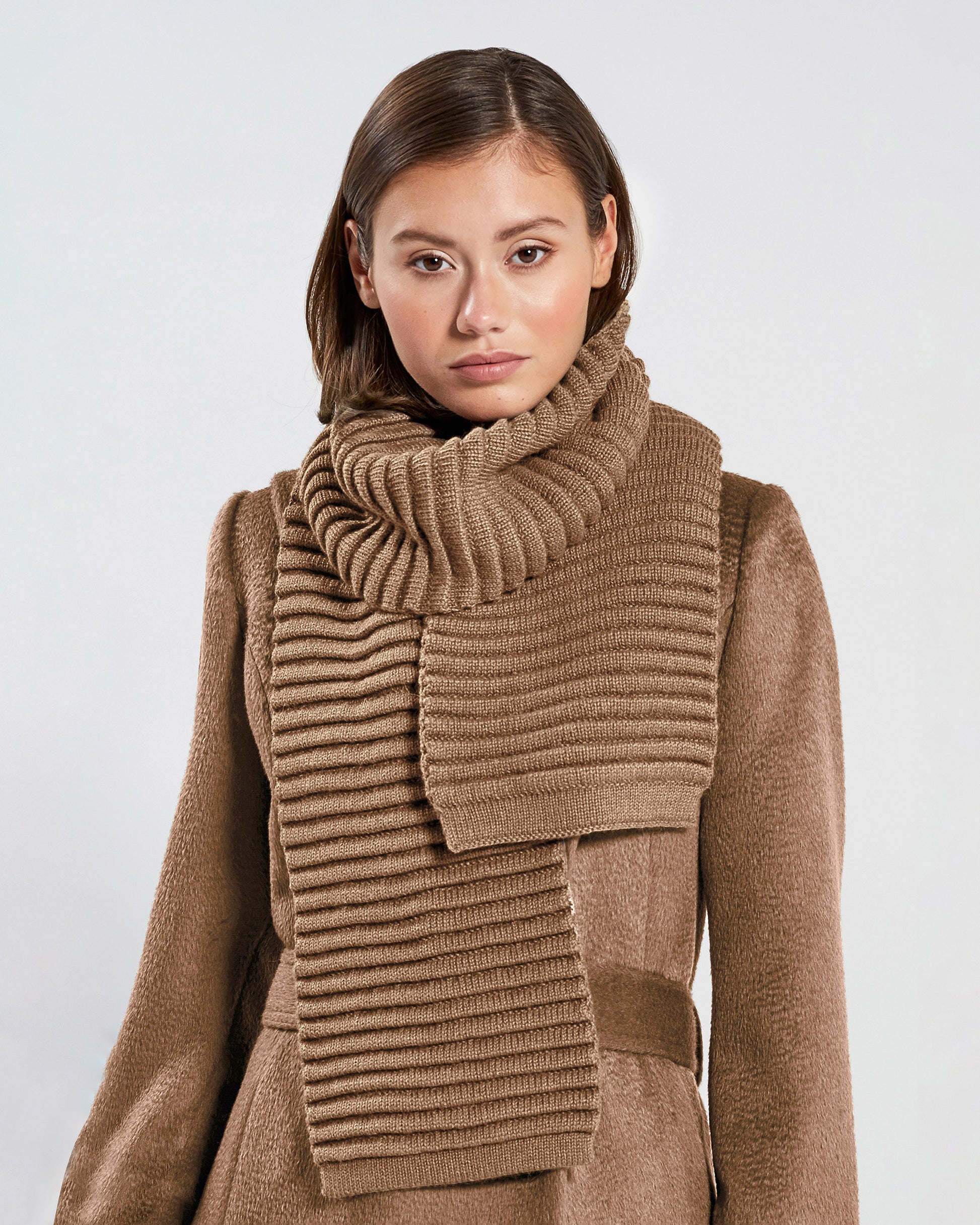 Front view of model wearing Sentaler's adult dark camel ribbed scarf, made of baby alpaca yarn, paired with a dark camel coat.