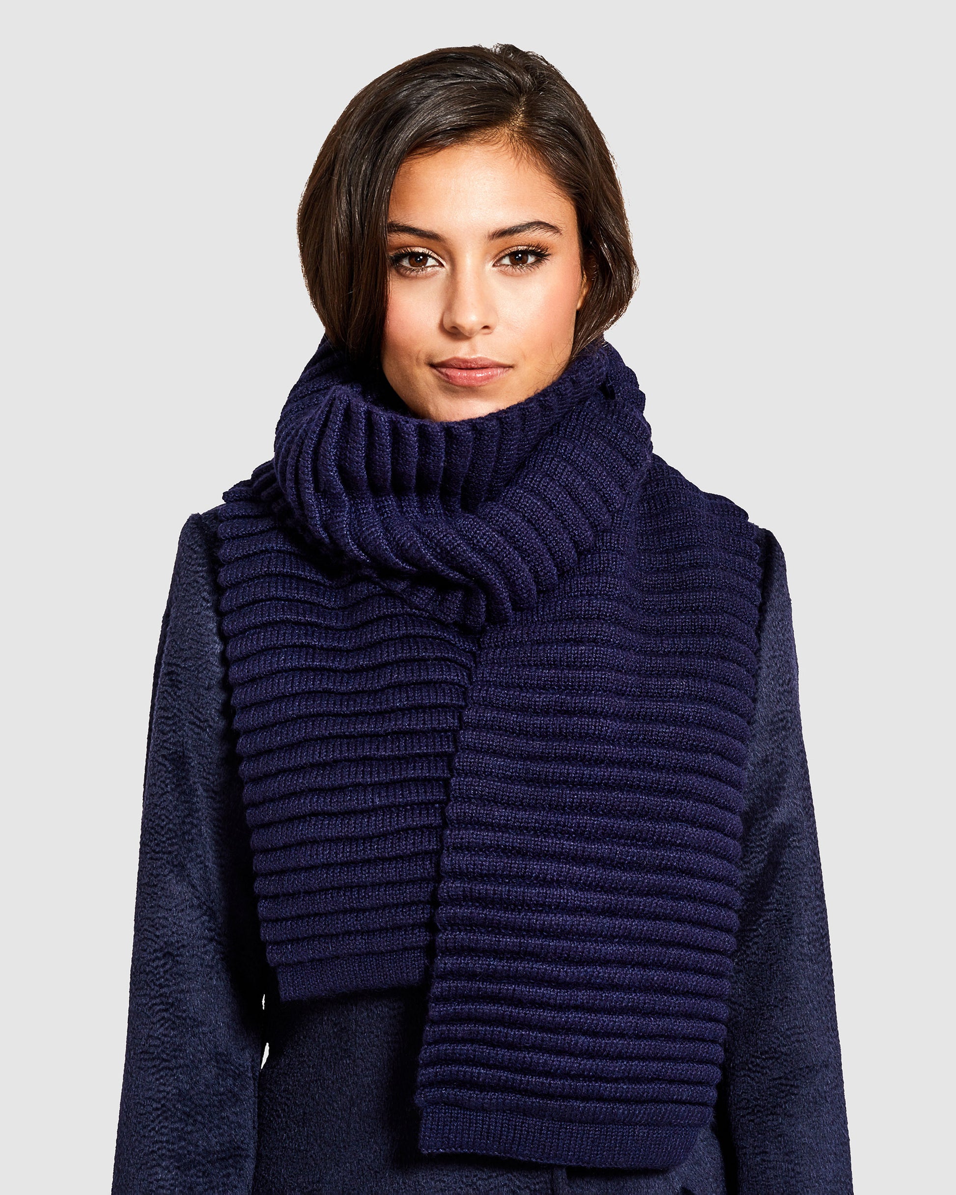 Front view of model wearing Sentaler's adult navy ribbed scarf, made of baby alpaca yarn, paired with a deep navy coat.