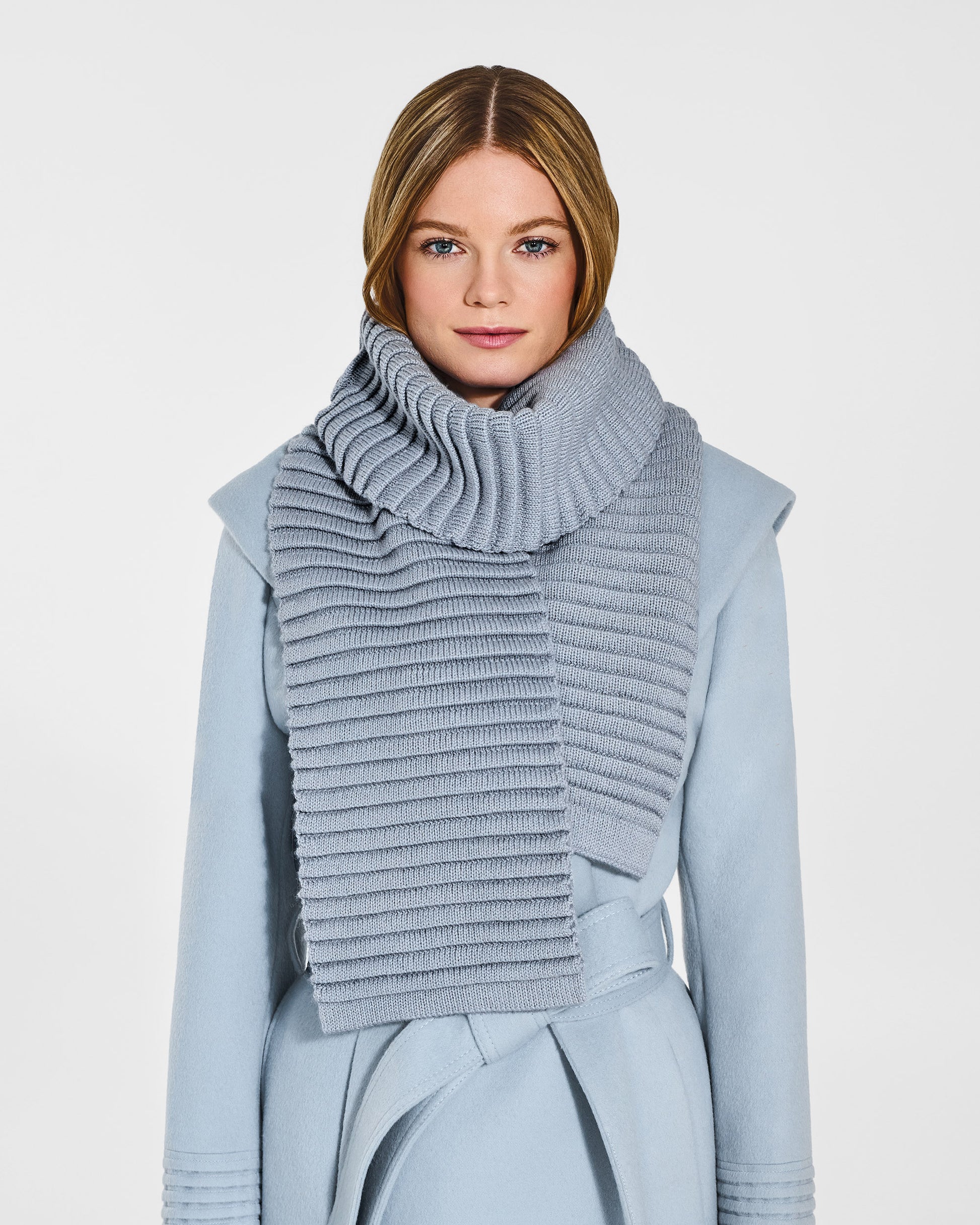 Front view of model wearing Sentaler's adult glacial blue ribbed scarf, made of baby alpaca yarn, paired with a glacial blue coat.