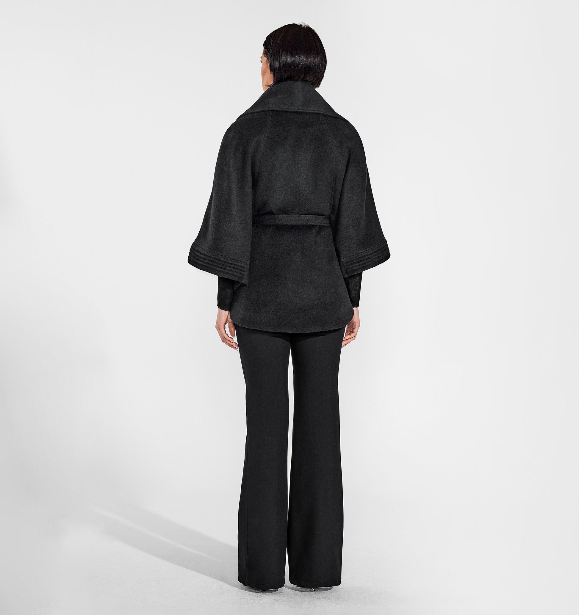 Back view of model wearing a Sentaler black cape made of baby alpaca wool, with a shawl collar, worn belted.