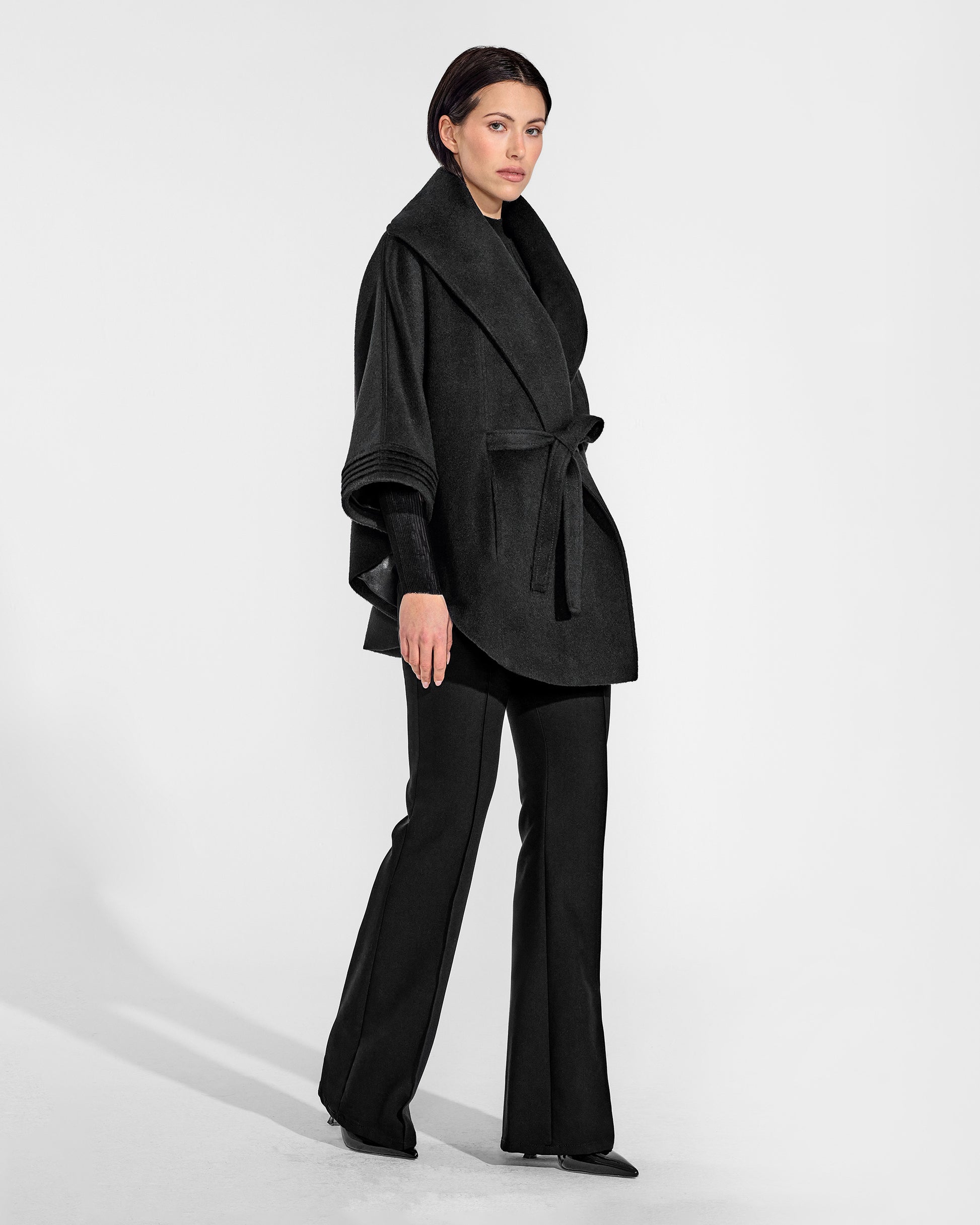 Side view of model wearing a Sentaler black cape made of baby alpaca wool, with a shawl collar, worn belted.