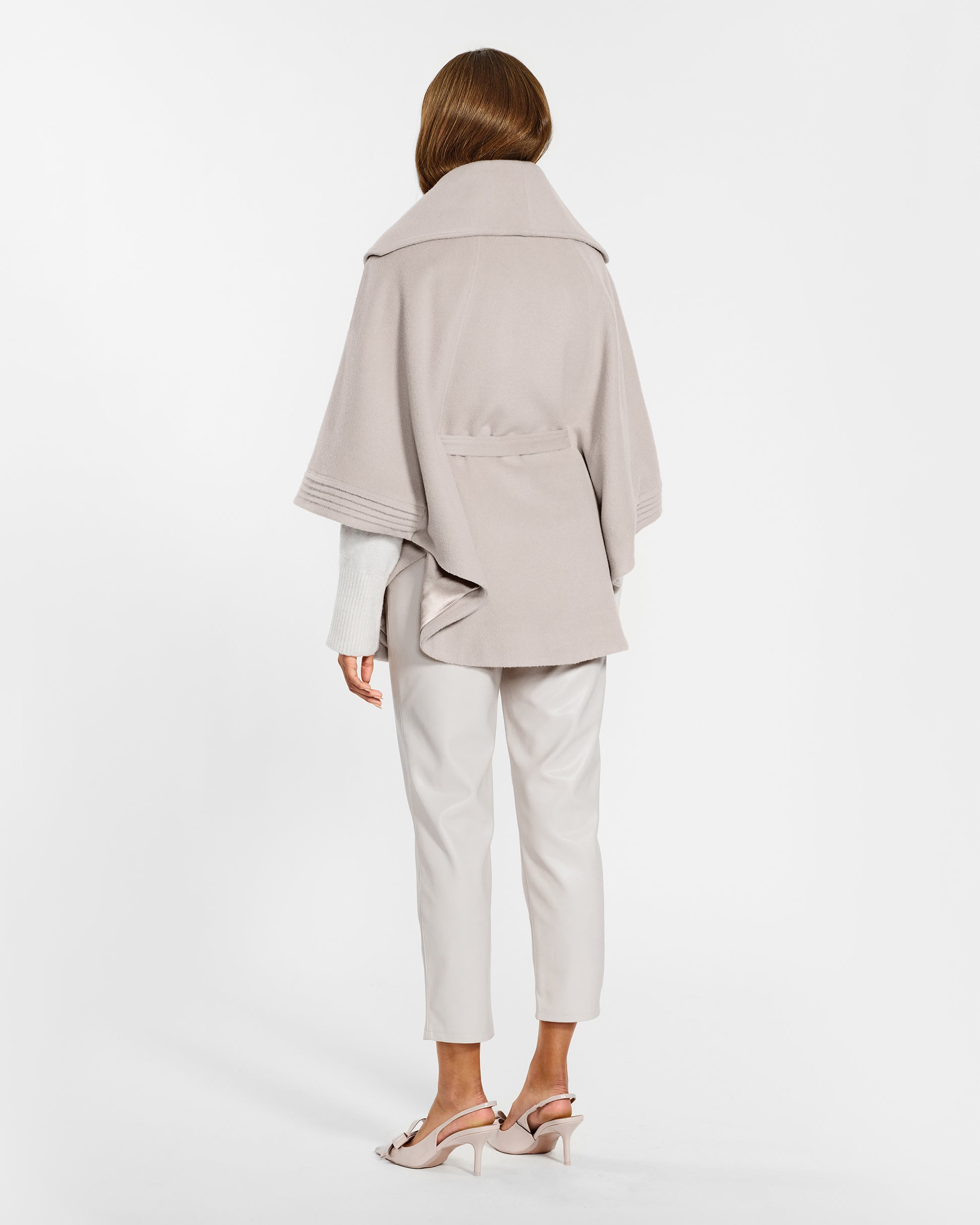 Back view of model wearing a Sentaler bleeker beige cape made of baby alpaca wool, with a shawl collar, worn belted.