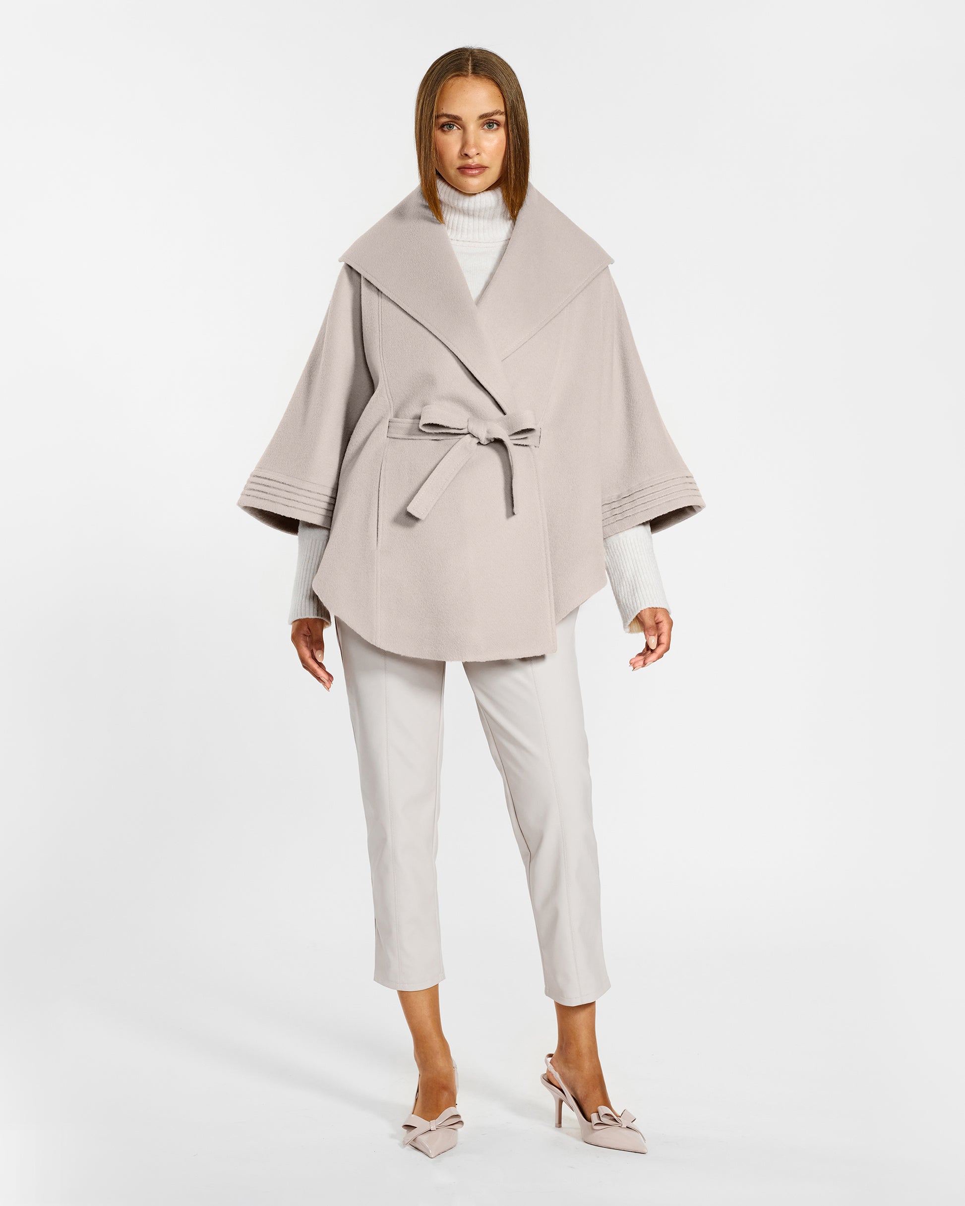 Front view of model wearing a Sentaler bleeker beige cape made of baby alpaca wool, with a shawl collar, worn belted.