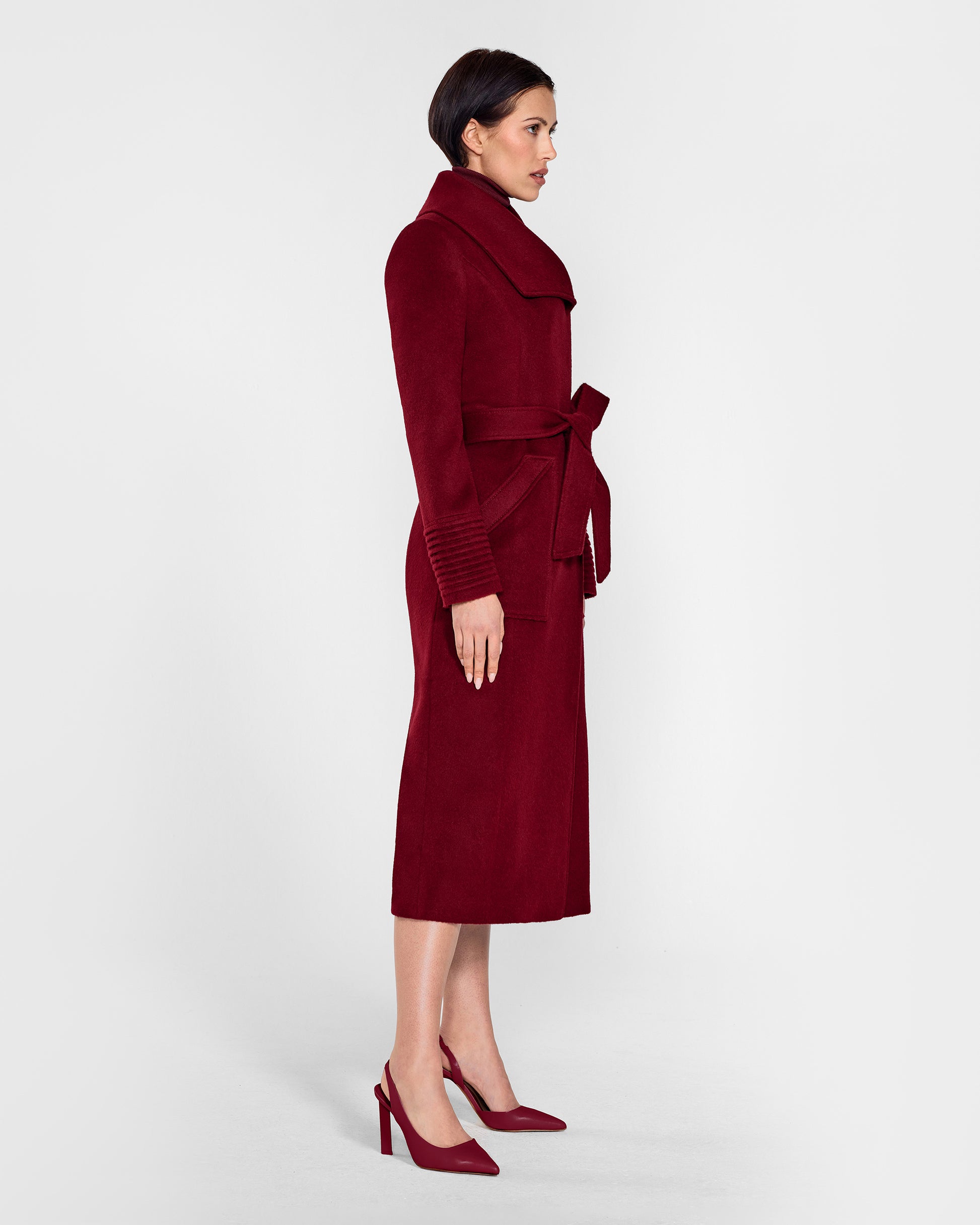 Side view of model wearing a Sentaler garnet red long wrap coat made of baby alpaca wool, with a wide collar, worn belted.