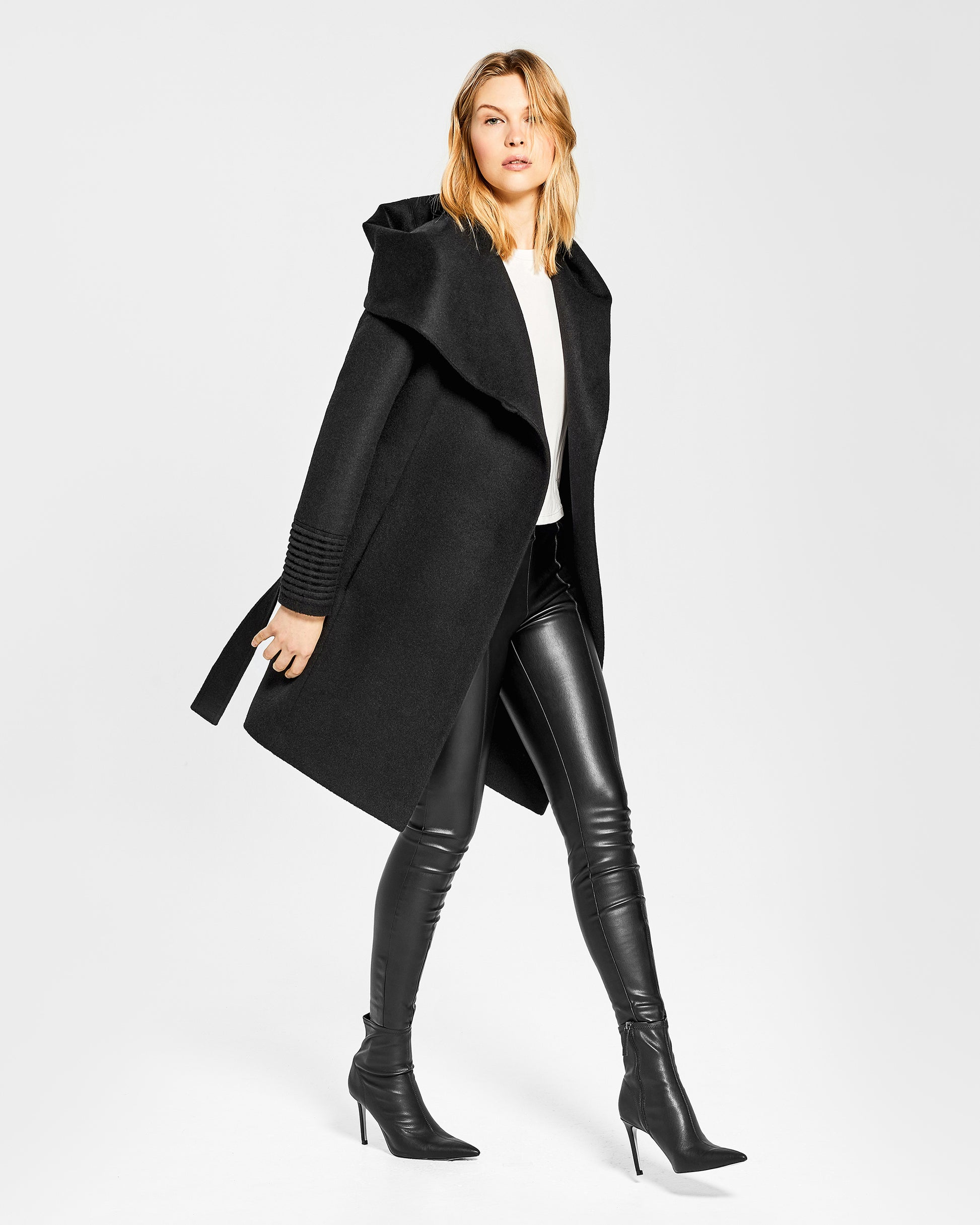 Side view of model wearing a Sentaler mid length black coat made of baby alpaca wool, with a hooded collar, worn open.