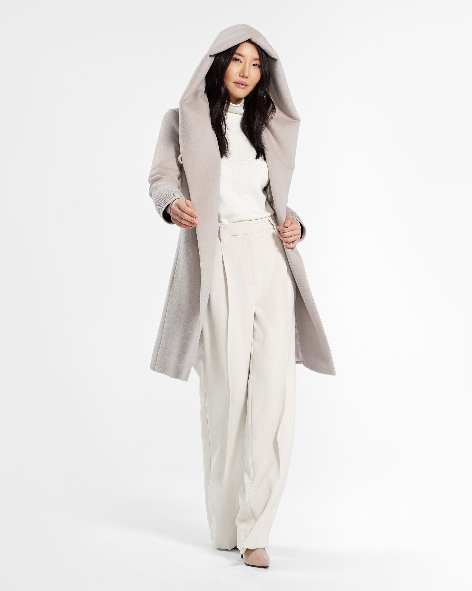 Front view of model wearing a Sentaler mid length bleeker beige coat made of baby alpaca wool, with a hooded collar, worn open.