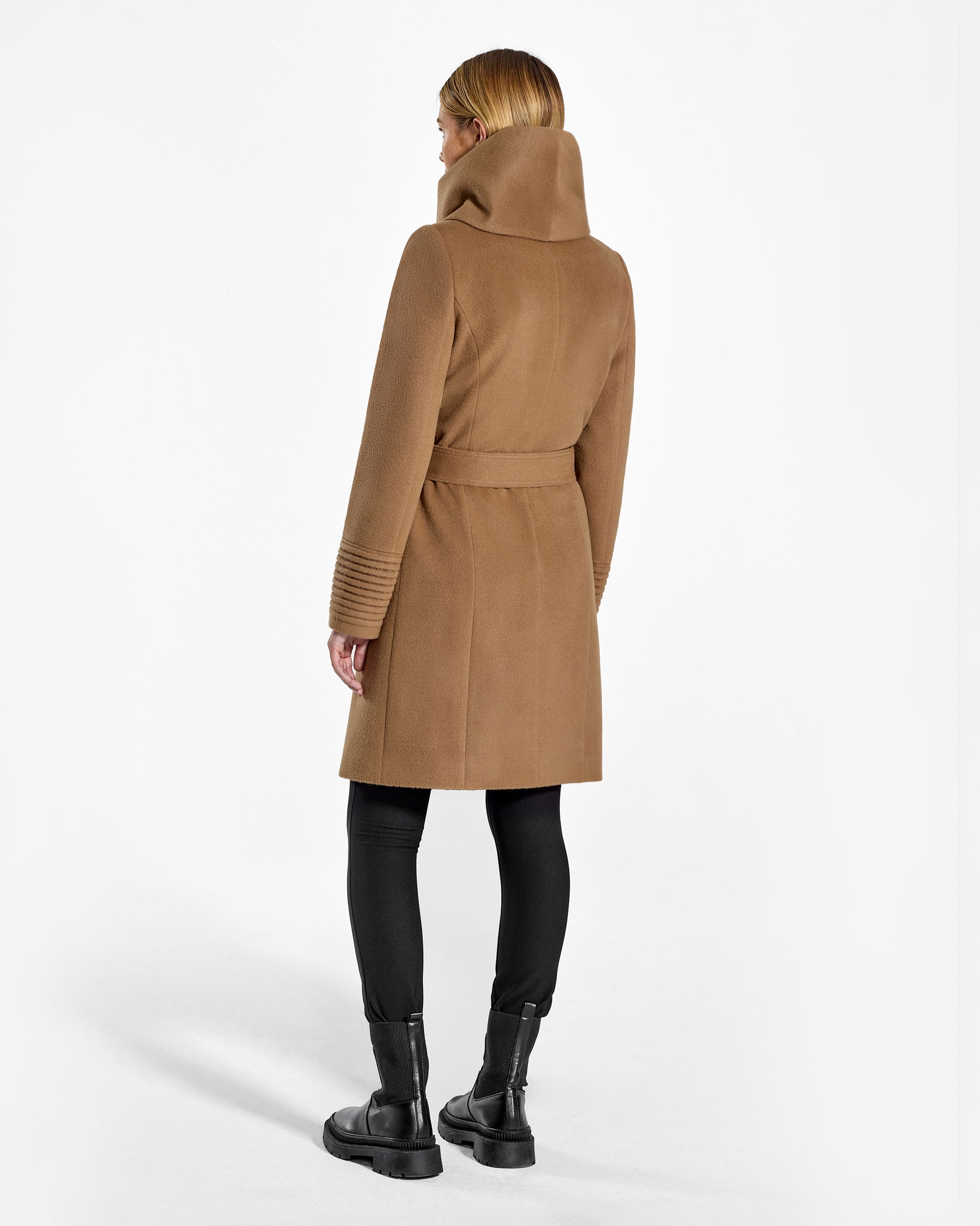 Back view of model wearing a Sentaler mid length dark camel coat made of baby alpaca wool, with a hooded collar, worn belted.