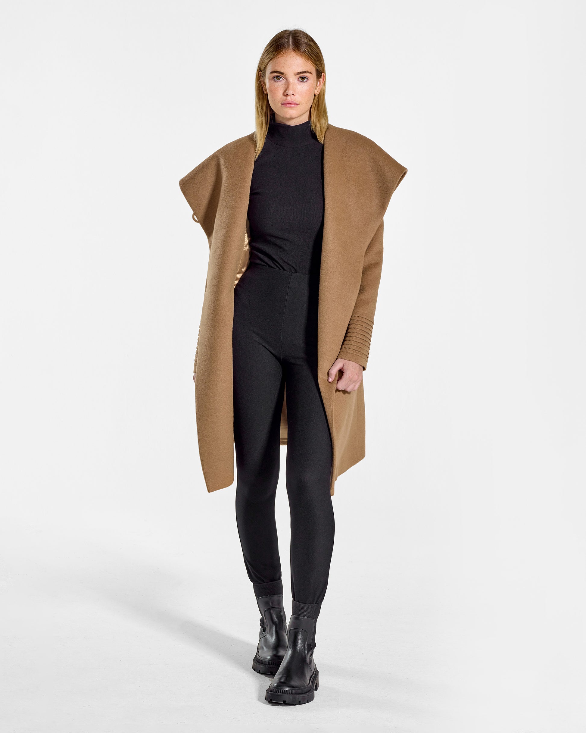 Front view of model wearing a Sentaler mid length dark camel coat made of baby alpaca wool, with a hooded collar, worn open.