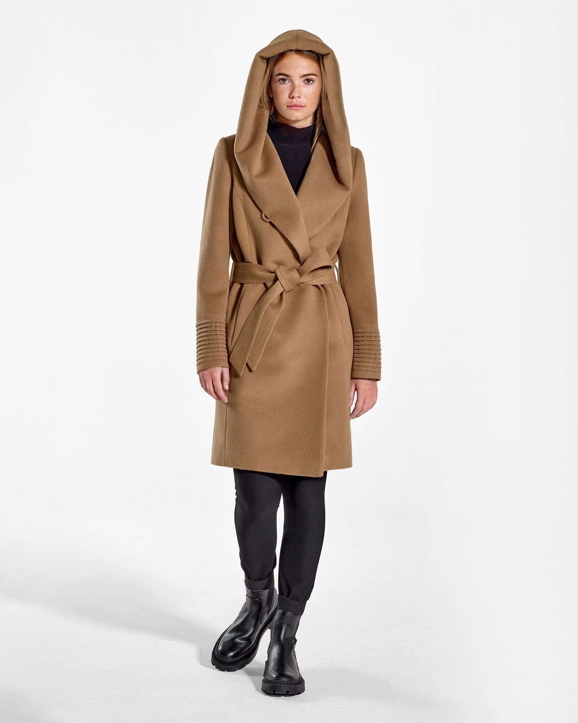 Front view of model wearing a Sentaler mid length dark camel coat made of baby alpaca wool, with a hooded collar, worn belted.