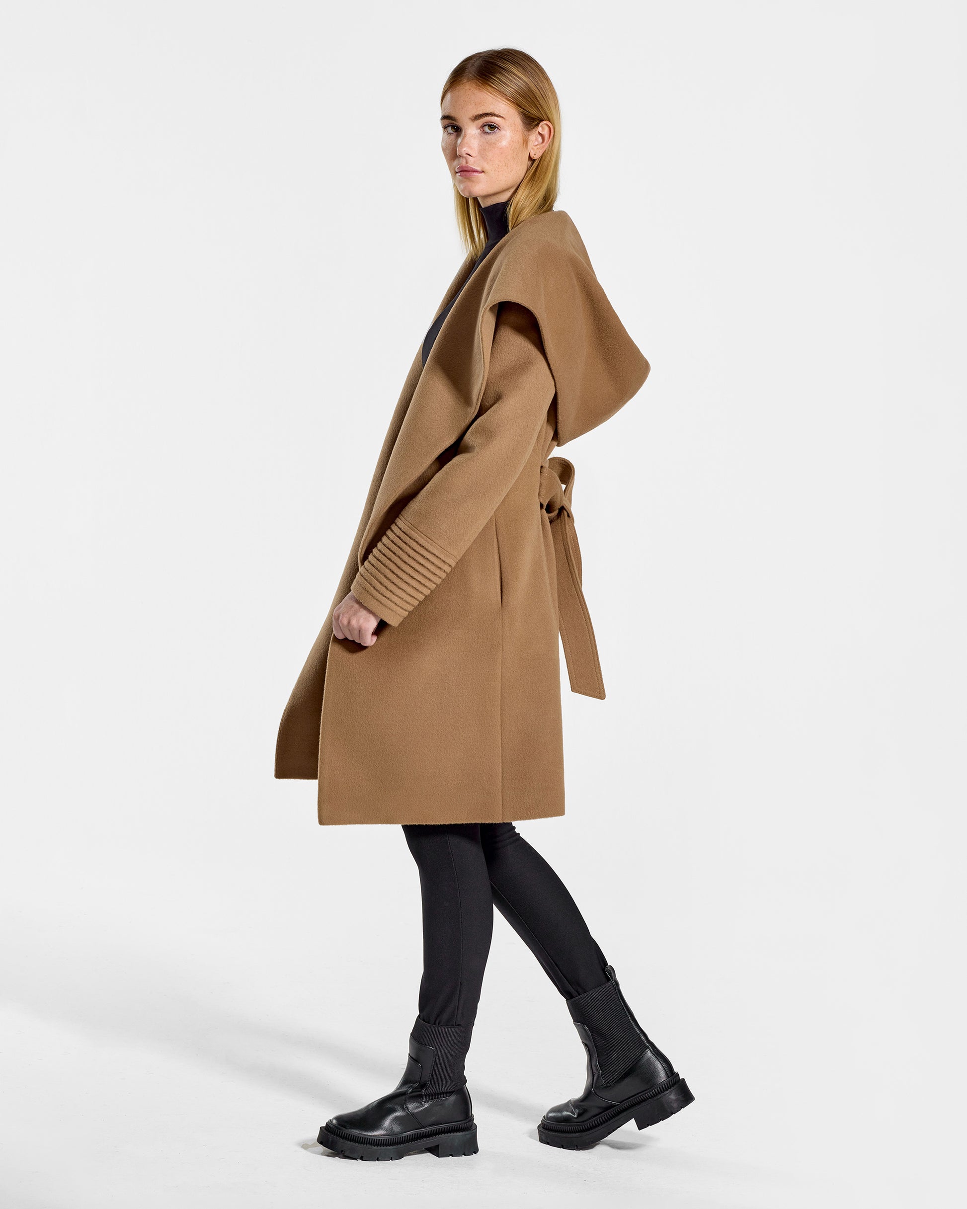 Side view of model wearing a Sentaler mid length dark camel coat made of baby alpaca wool, with a hooded collar, worn open.