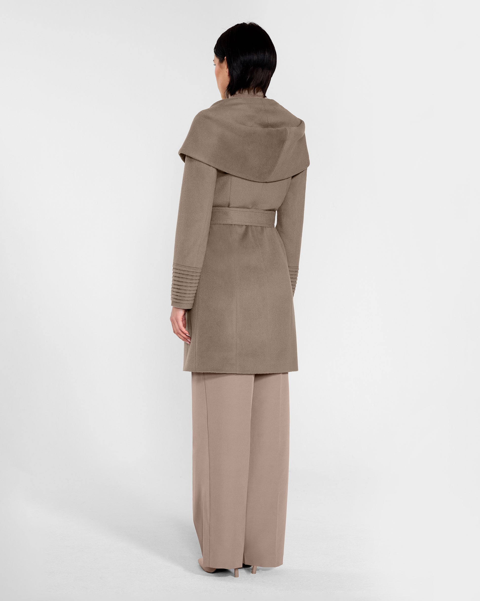Back view of model wearing a Sentaler mid length warm taupe coat made of baby alpaca wool, with a hooded collar, worn belted.