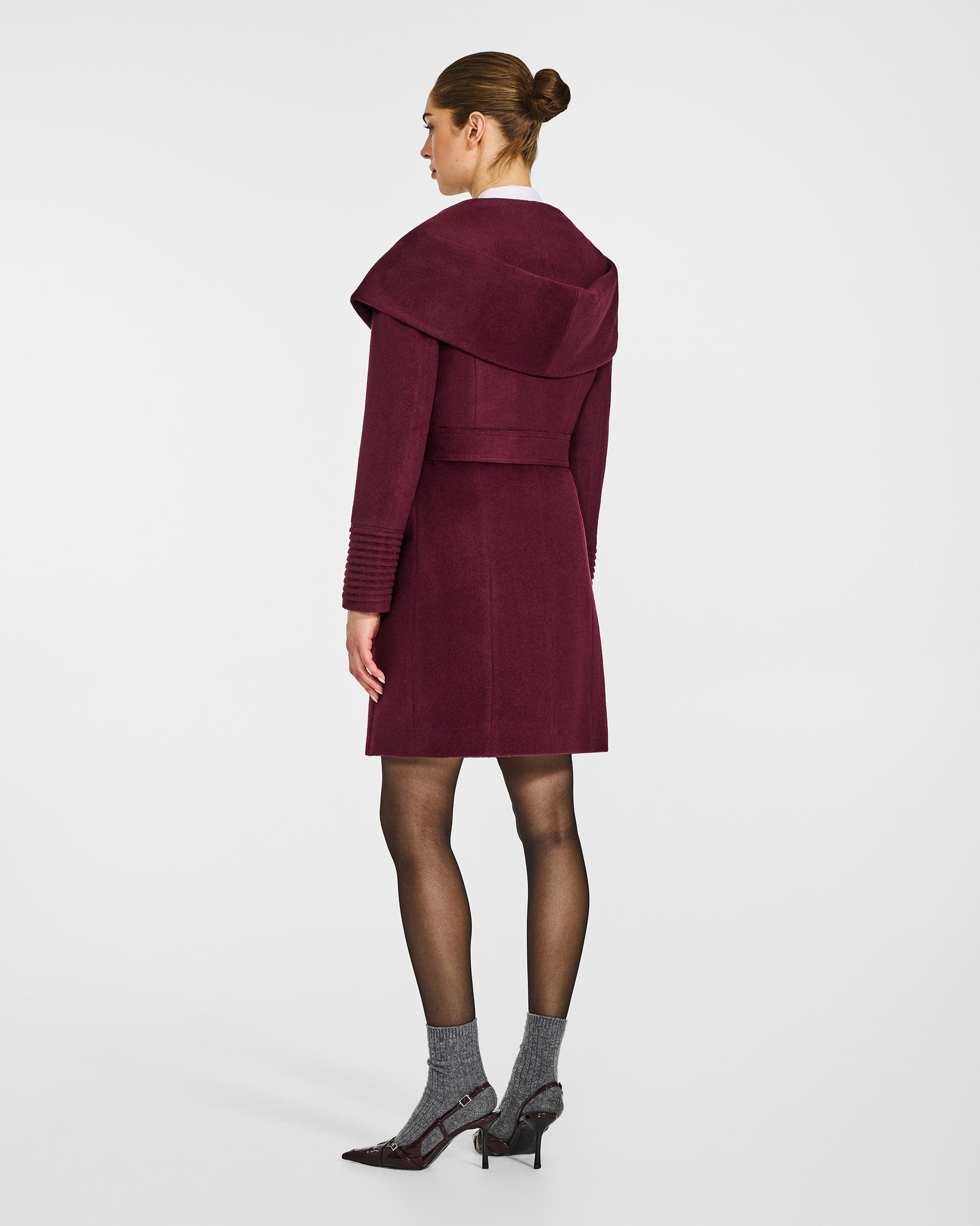 Back view of model wearing a Sentaler mid length cherry lacquer coat made of baby alpaca wool, with a hooded collar, worn belted with shawl collar.