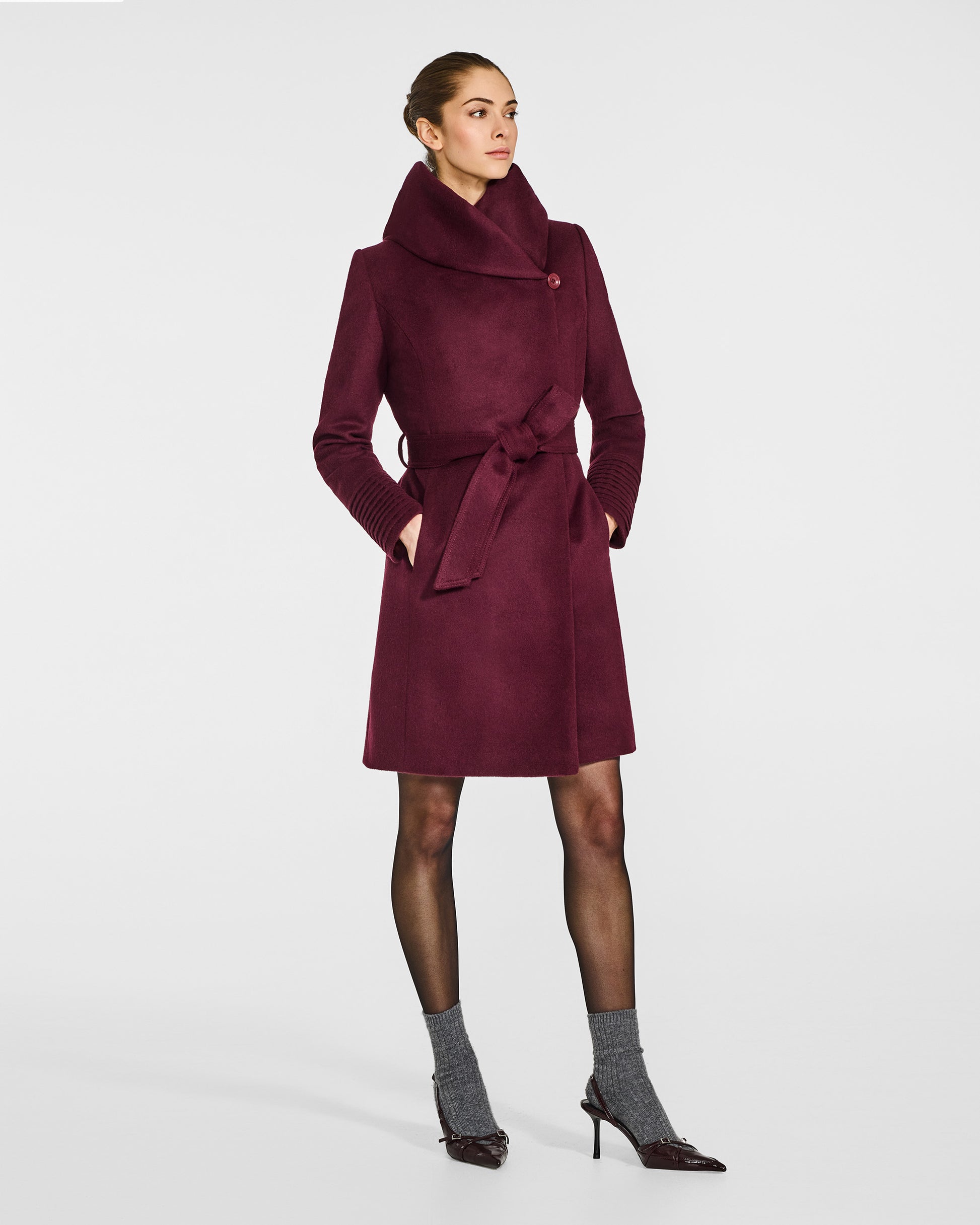 Side view of model wearing a Sentaler mid length cherry lacquer coat made of baby alpaca wool, with a hooded collar, worn belted.