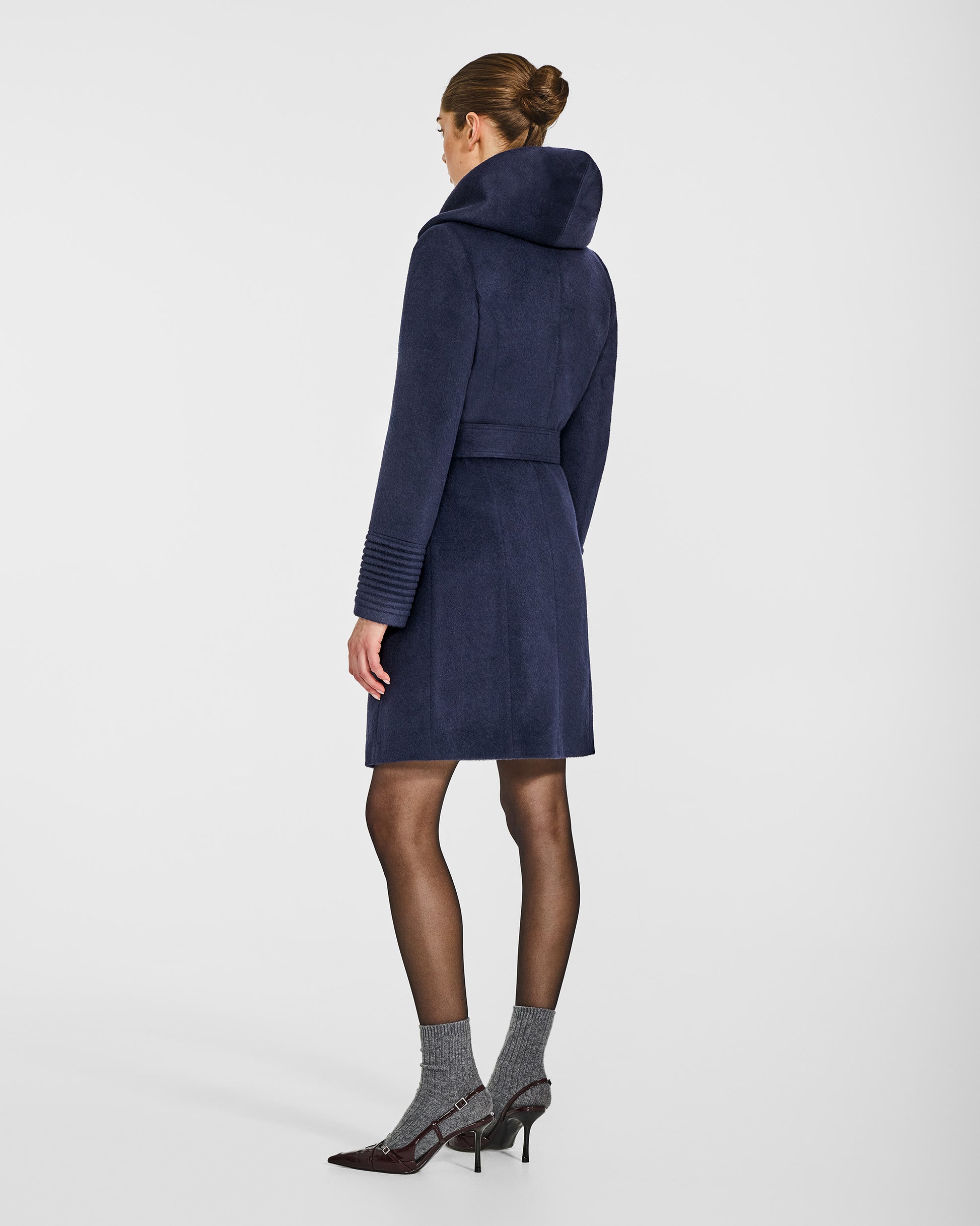 Back view of model wearing a Sentaler mid length deep navy coat made of baby alpaca wool, with a hooded collar, worn belted.