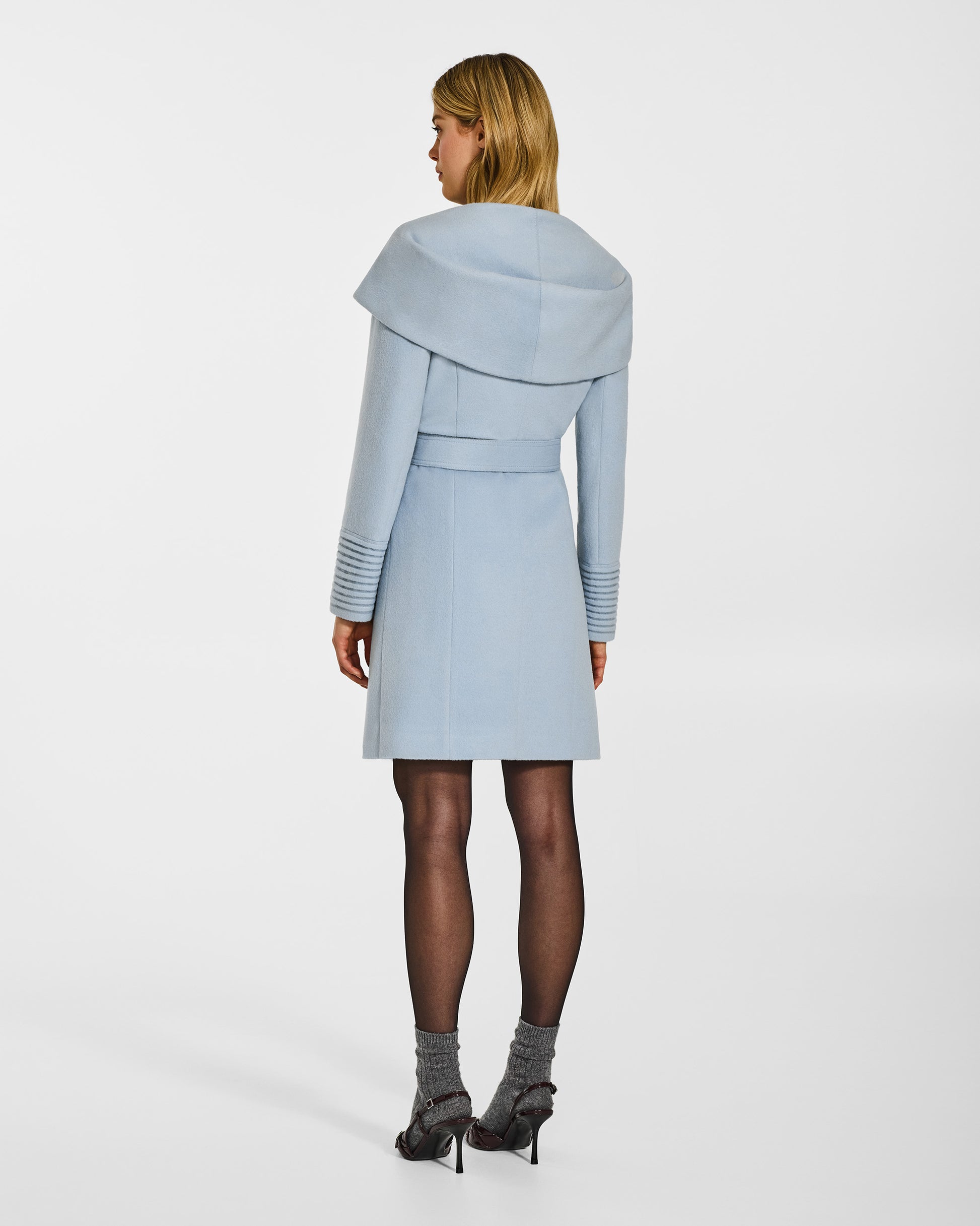 Back view of model wearing a Sentaler mid length glacial blue coat made of baby alpaca wool, with a hooded collar, worn belted.
