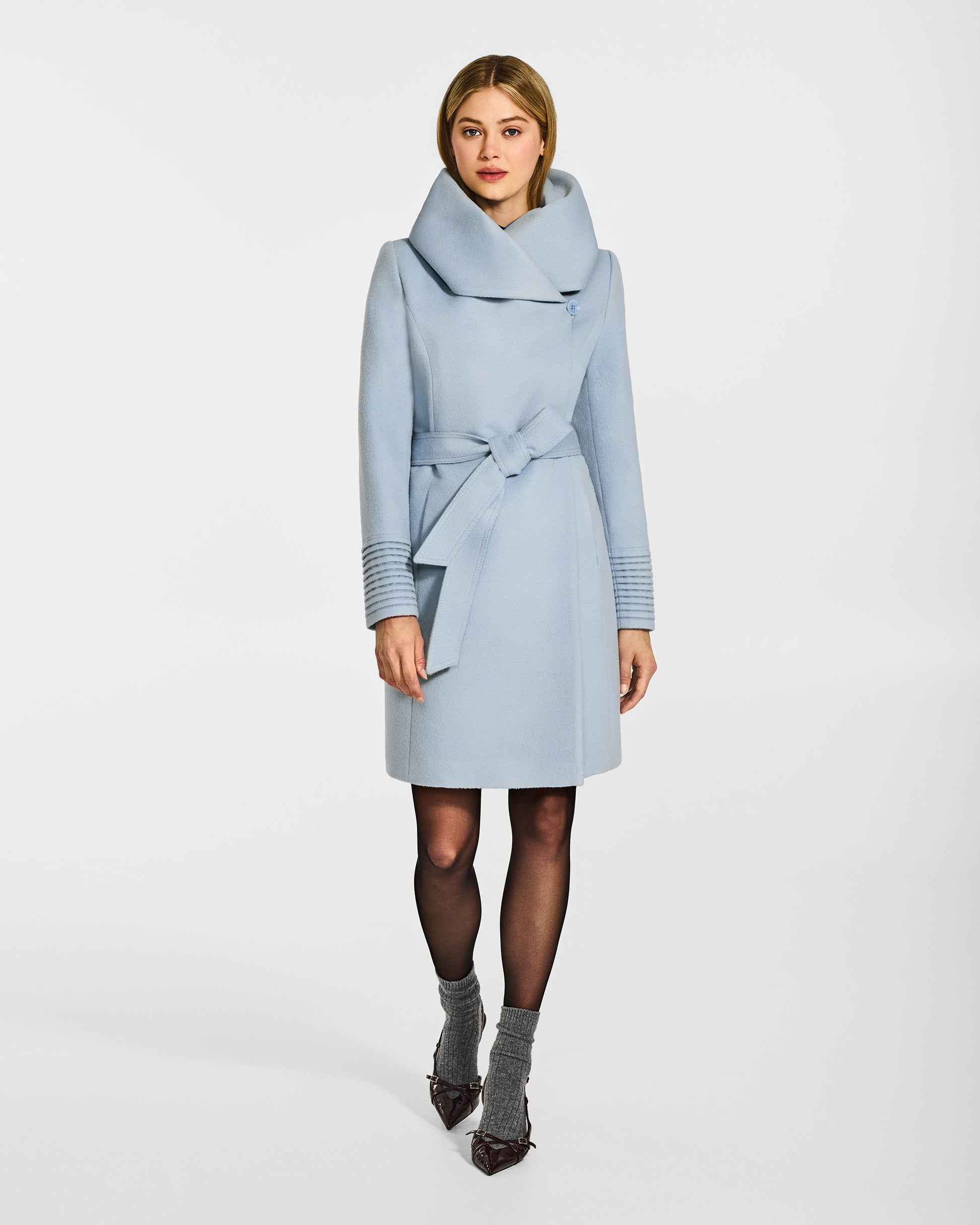 Front view of model wearing a Sentaler mid length glacial blue coat made of baby alpaca wool, with a hooded collar, worn belted.
