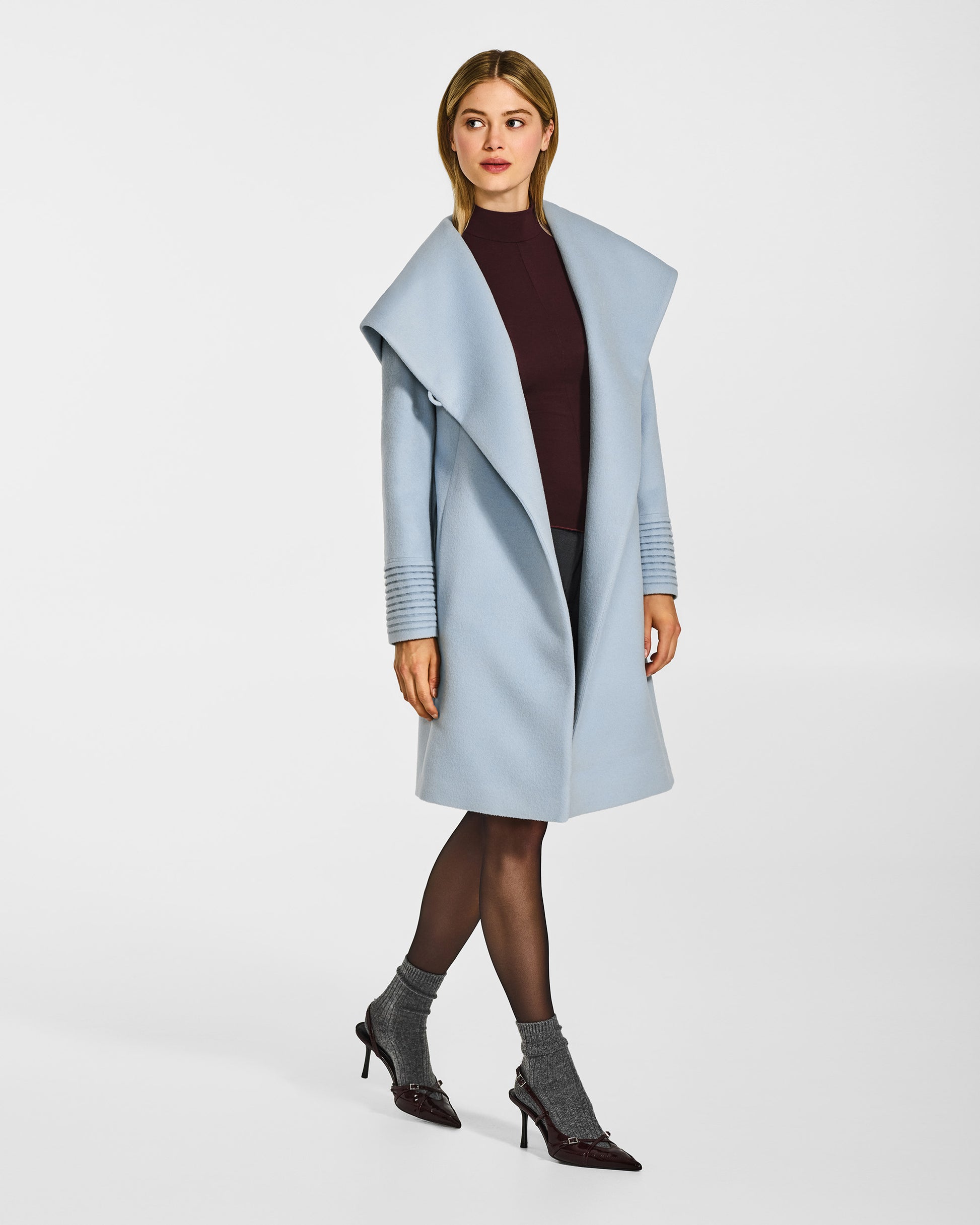 Side view of model wearing a Sentaler mid length glacial blue coat made of baby alpaca wool, with a hooded collar, worn open.