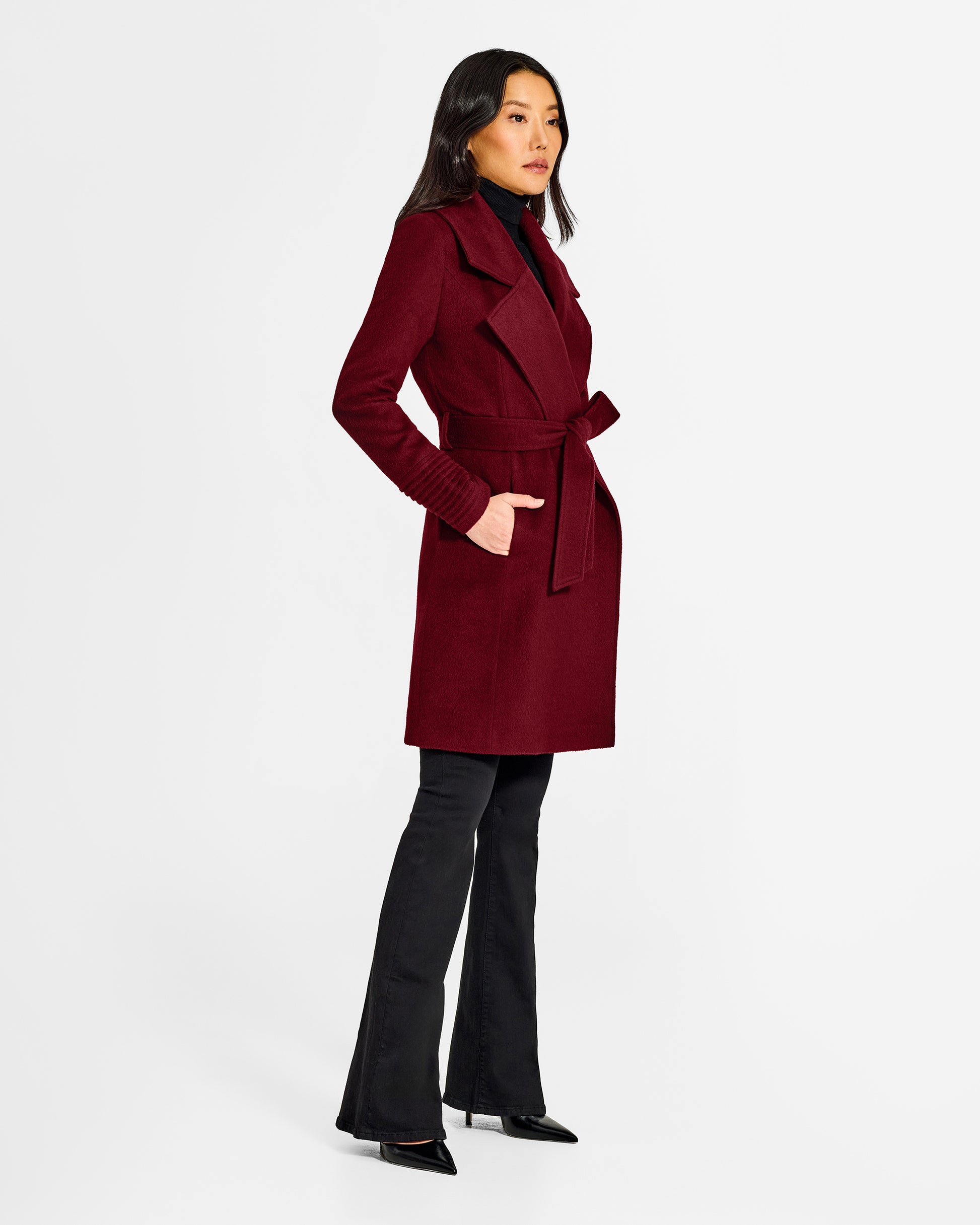 Side view of model wearing a Sentaler mid length garnet red coat made of baby alpaca wool, with a large notched collar, worn belted.