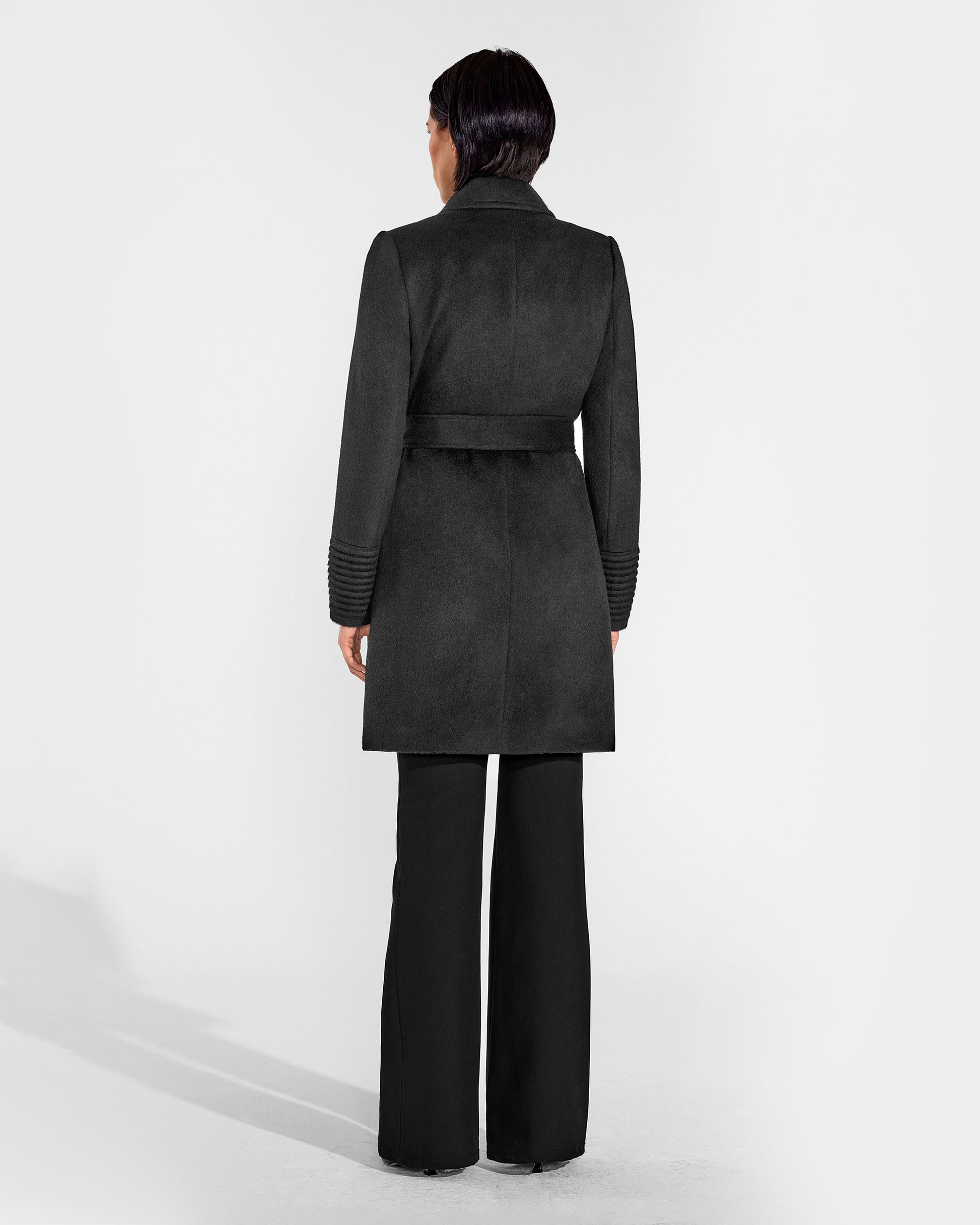 Back view of model wearing a Sentaler mid length black coat made of baby alpaca wool, with a notched collar, worn belted.