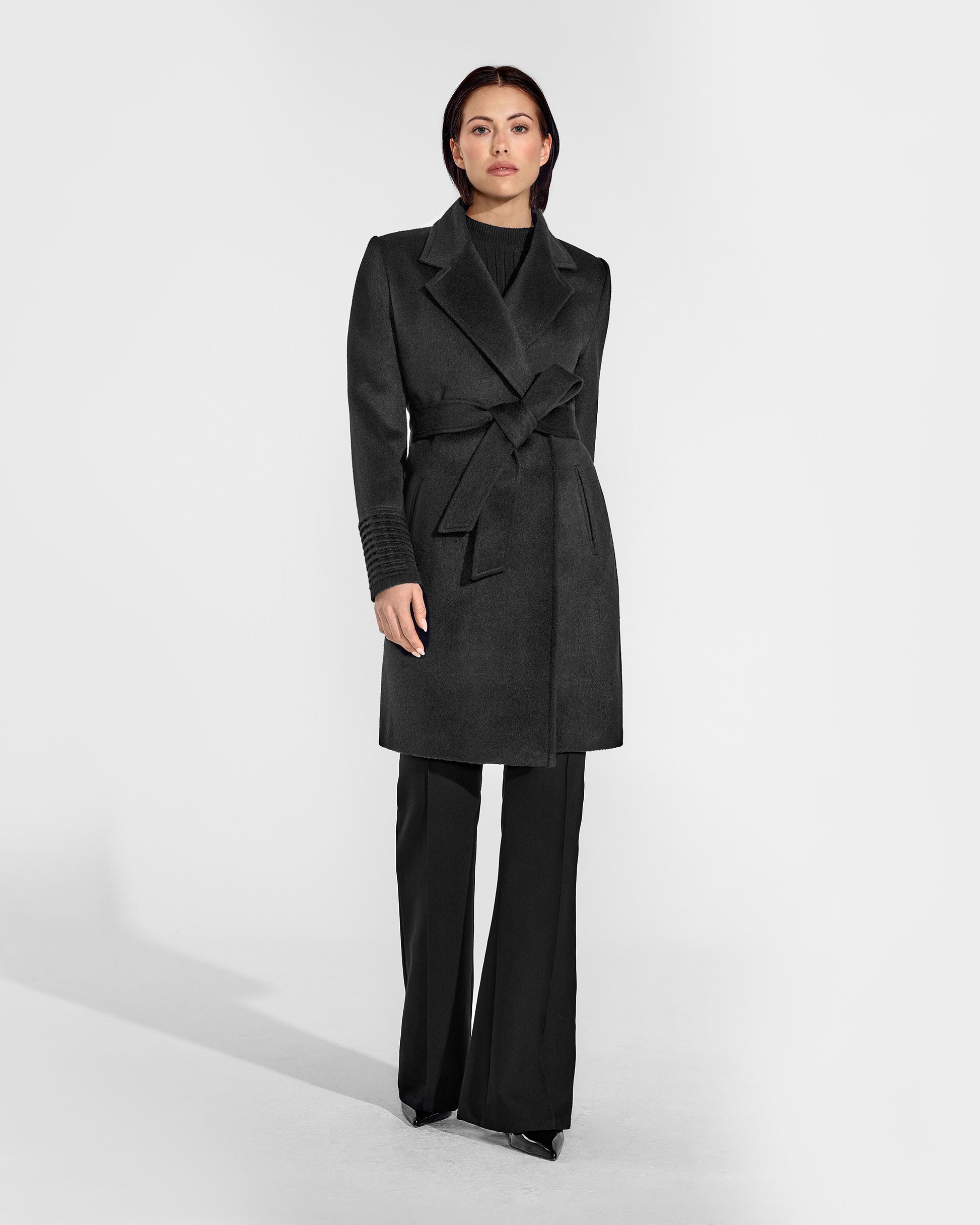 Front view of model wearing a Sentaler mid length black coat made of baby alpaca wool, with a notched collar, worn belted.