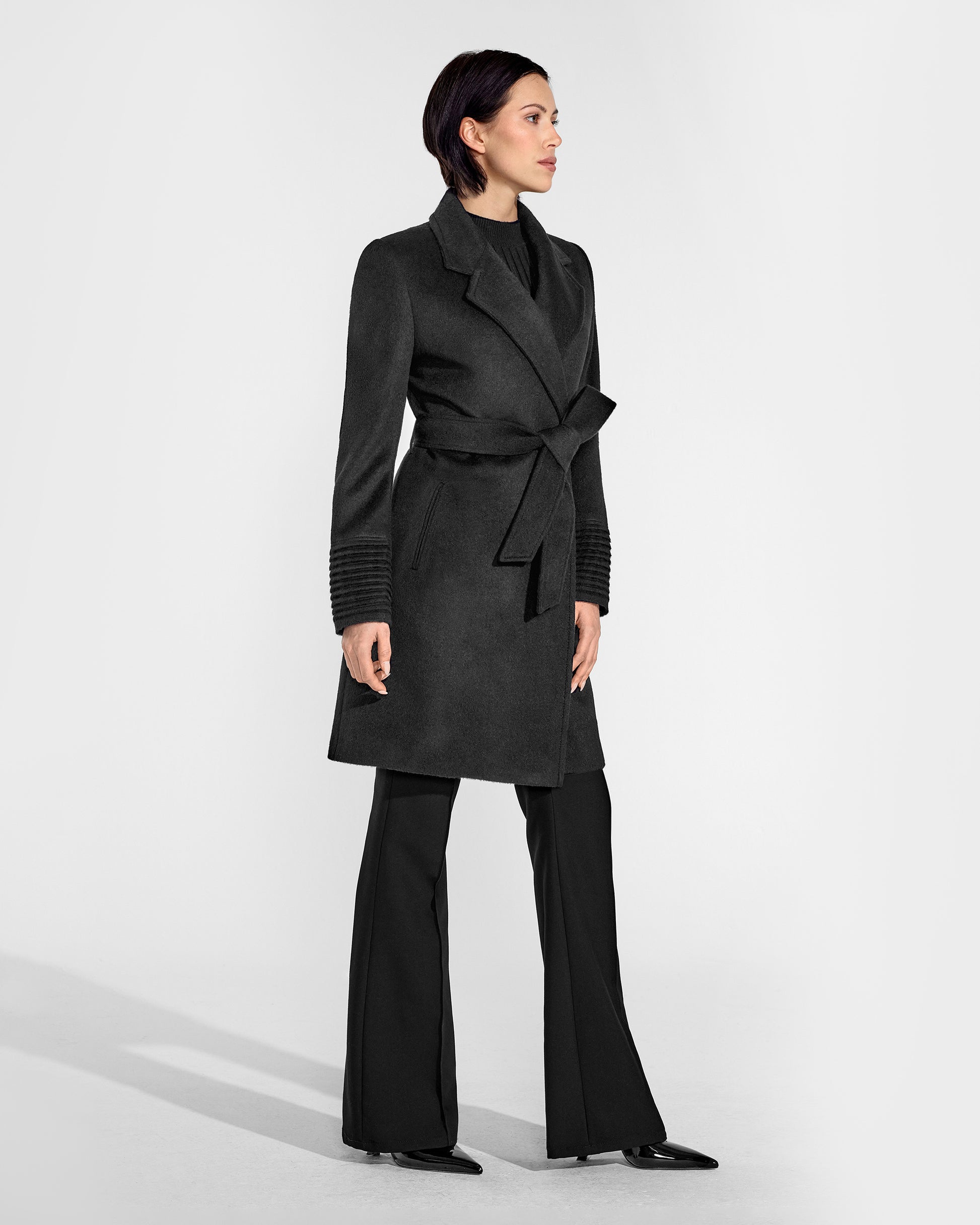 Side view of model wearing a Sentaler mid length black coat made of baby alpaca wool, with a notched collar, worn belted.