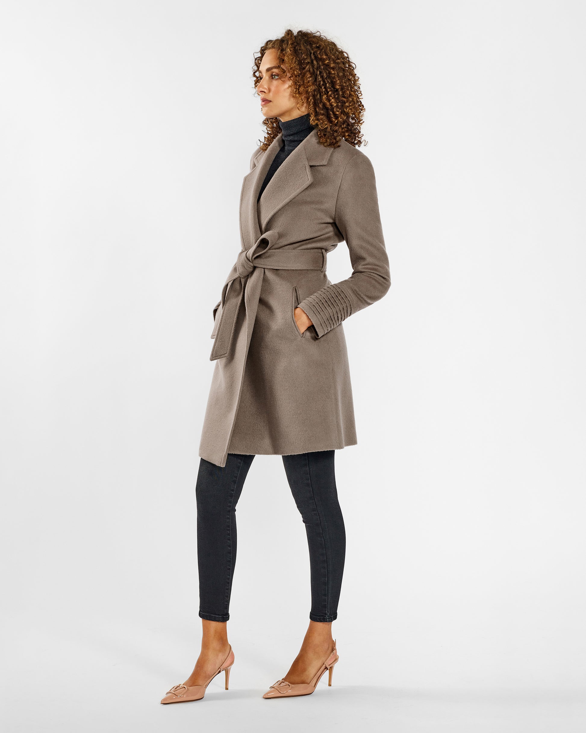 Side view of model wearing a Sentaler mid length warm taupe coat made of baby alpaca wool, with a notched collar, worn belted.