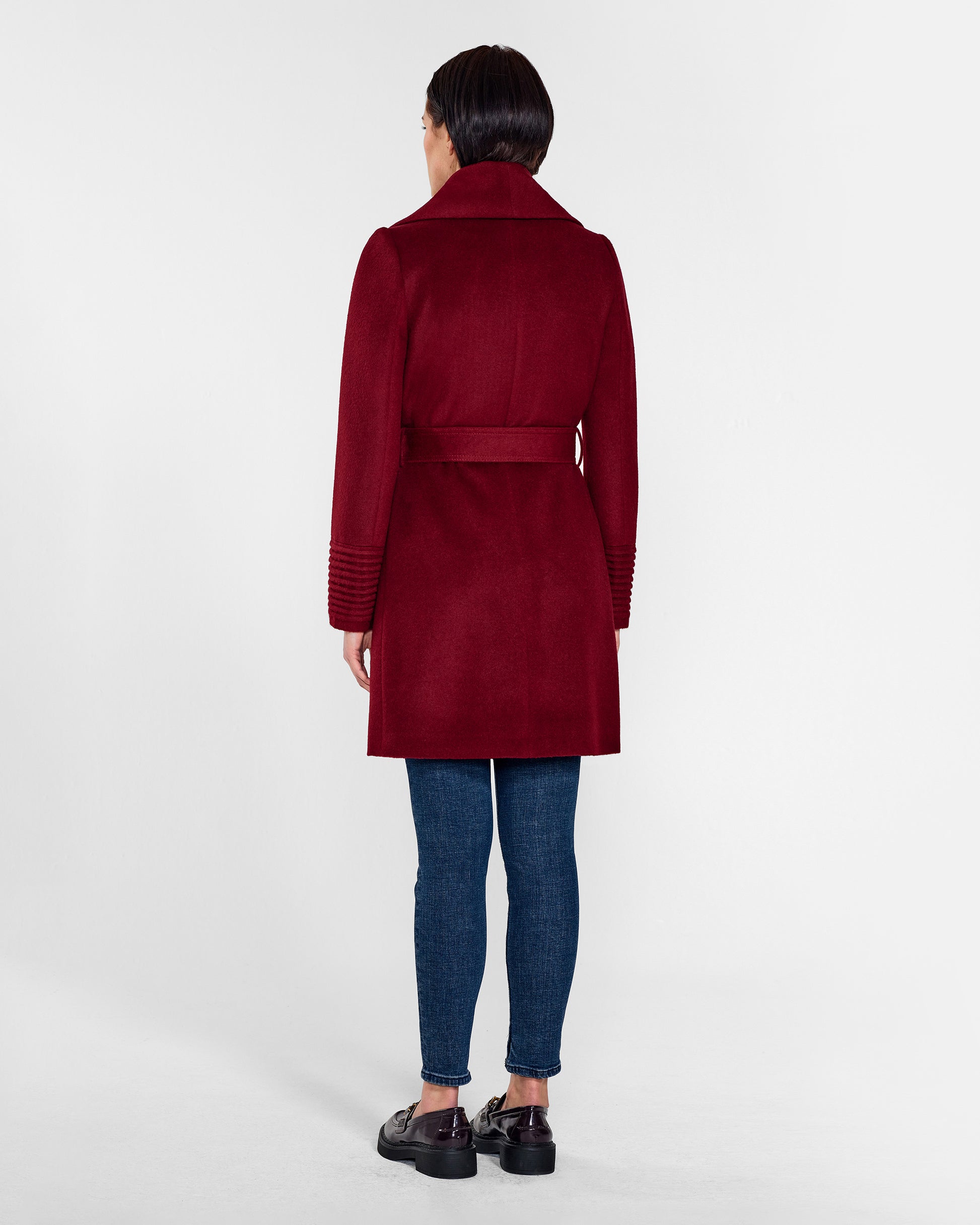 Back view of model wearing a Sentaler mid length garnet red coat made of baby alpaca wool, with a shawl collar, worn belted.