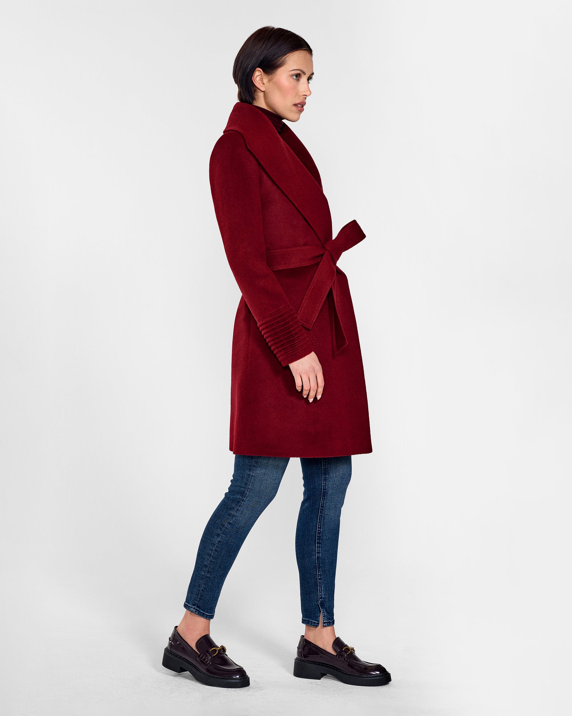 Side view of model wearing a Sentaler mid length garnet red coat made of baby alpaca wool, with a shawl collar, worn belted.
