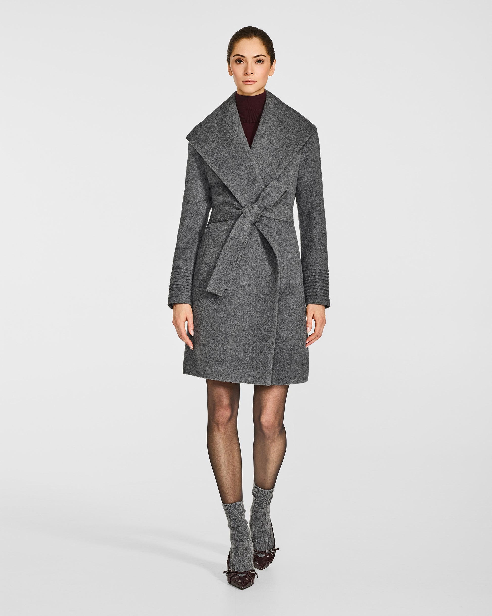 Front view of model wearing a Sentaler mid length grey mélange coat made of baby alpaca wool, with a shawl collar, worn belted.