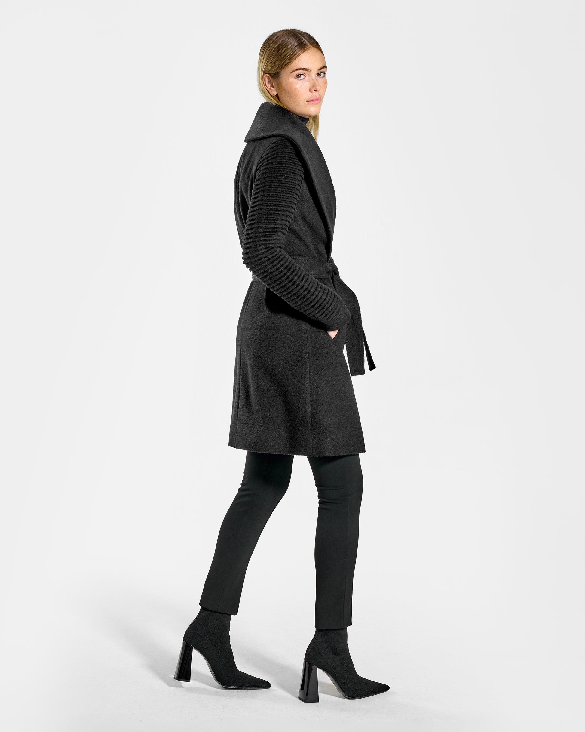 Side view of model wearing a Sentaler mid length black wrap coat made of baby alpaca wool, with ribbed sleeves, worn belted.