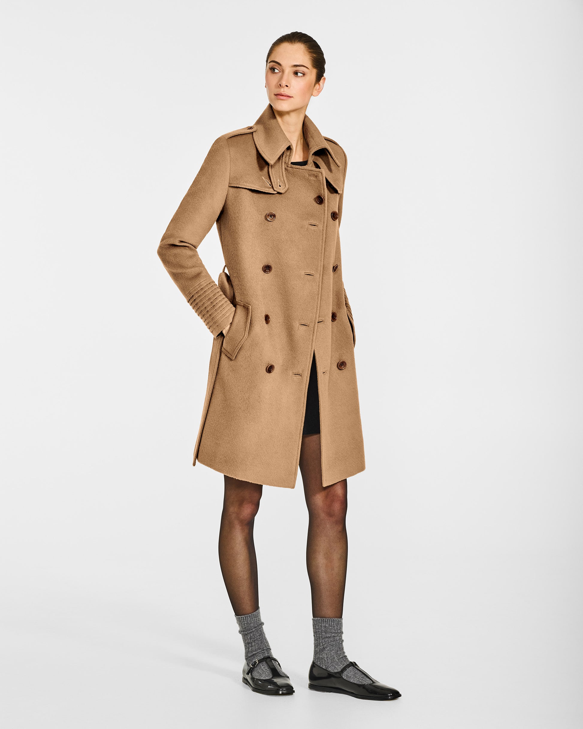 Side view of model wearing a Sentaler mid length dark camel trench coat made of baby alpaca wool, with a spread collar, worn partially buttoned.