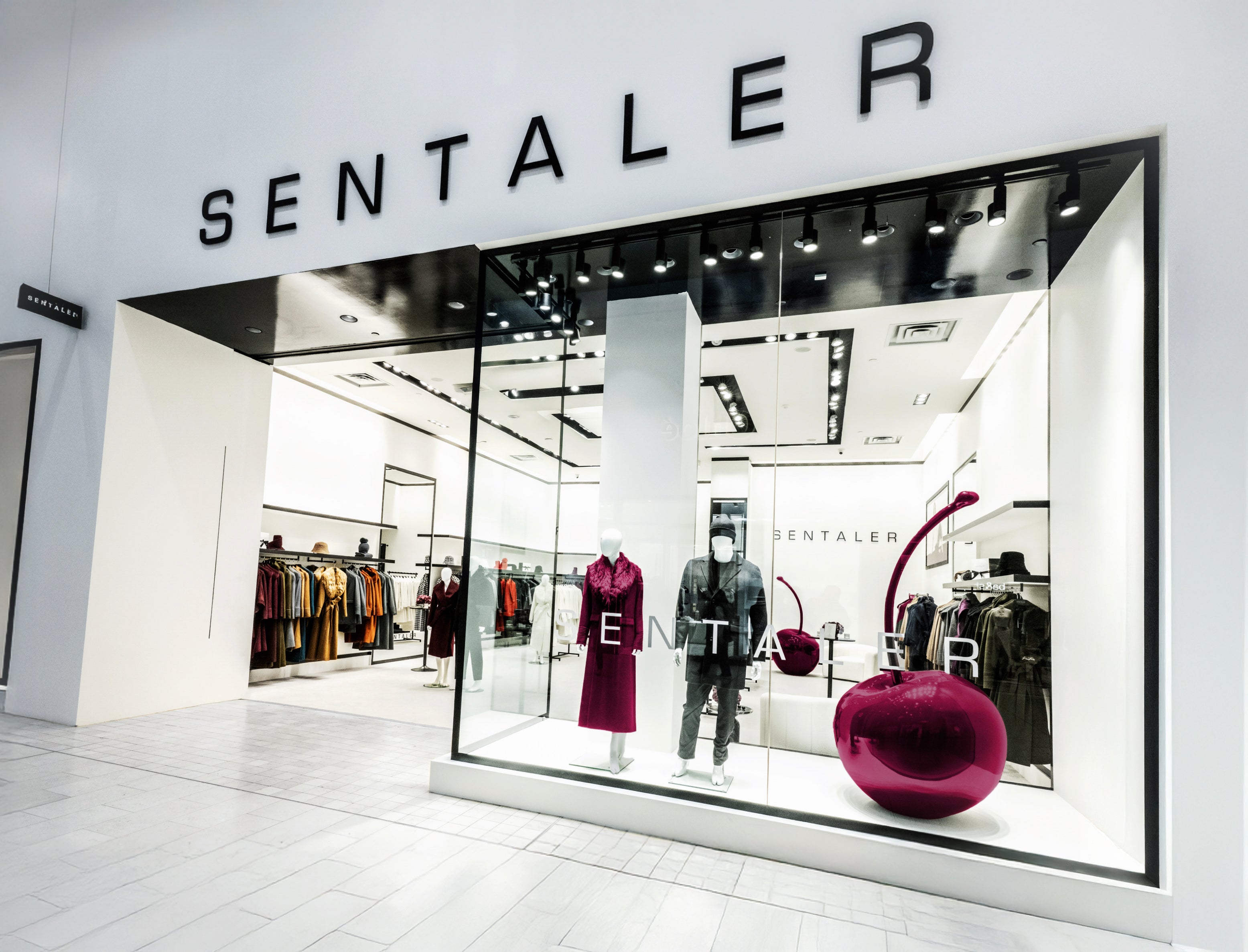 Exterior view of the SENTALER store at Yorkdale Shopping Centre featuring a sleek modern design with black and white décor, mannequins dressed in stylish coats, and a large glossy pink cherry sculpture in the display window.