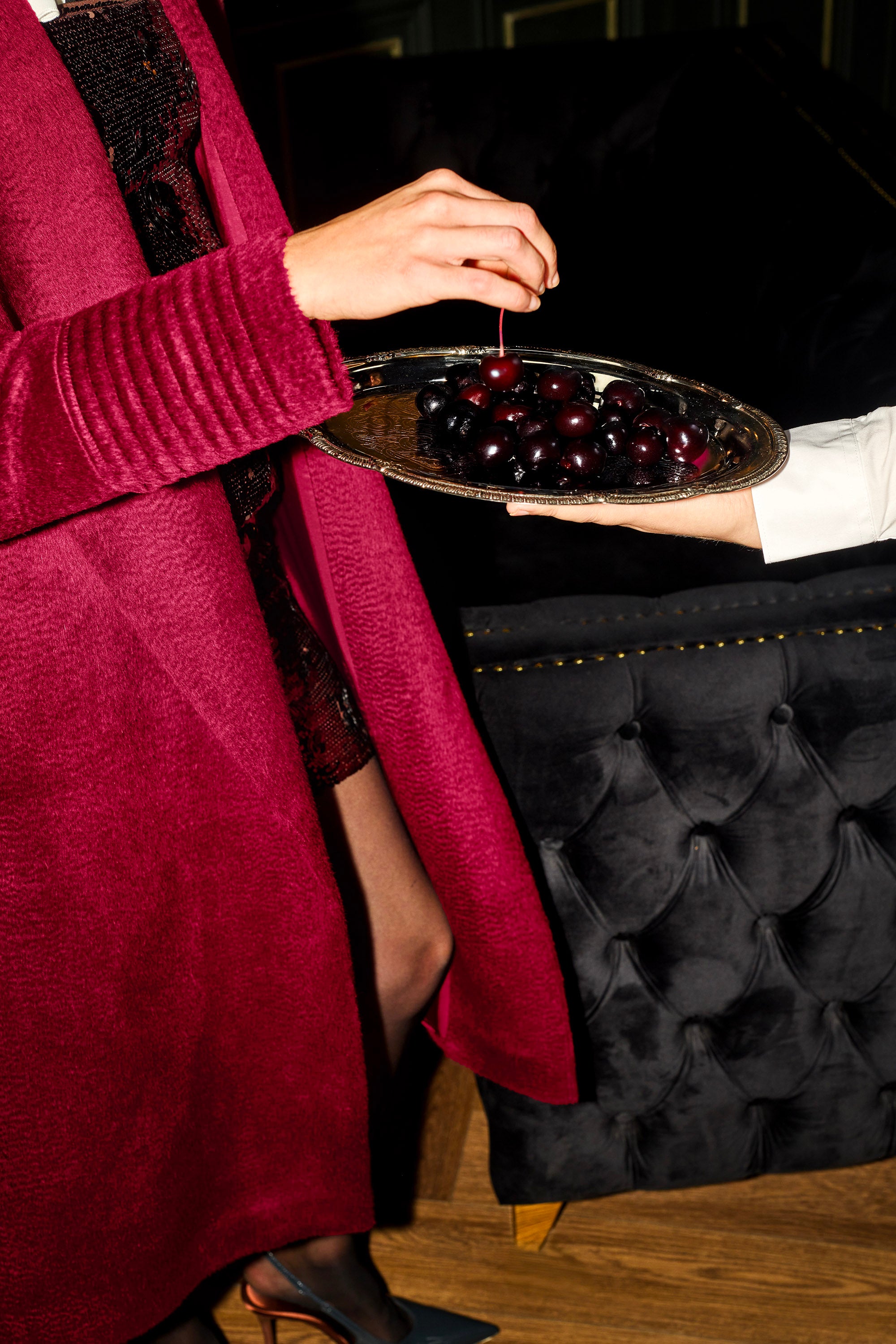 Side view of model wearing a Sentaler long bordeaux wrap coat made of suri alpaca wool, with a shawl collar, worn open while picking up cherries from a silver platter.