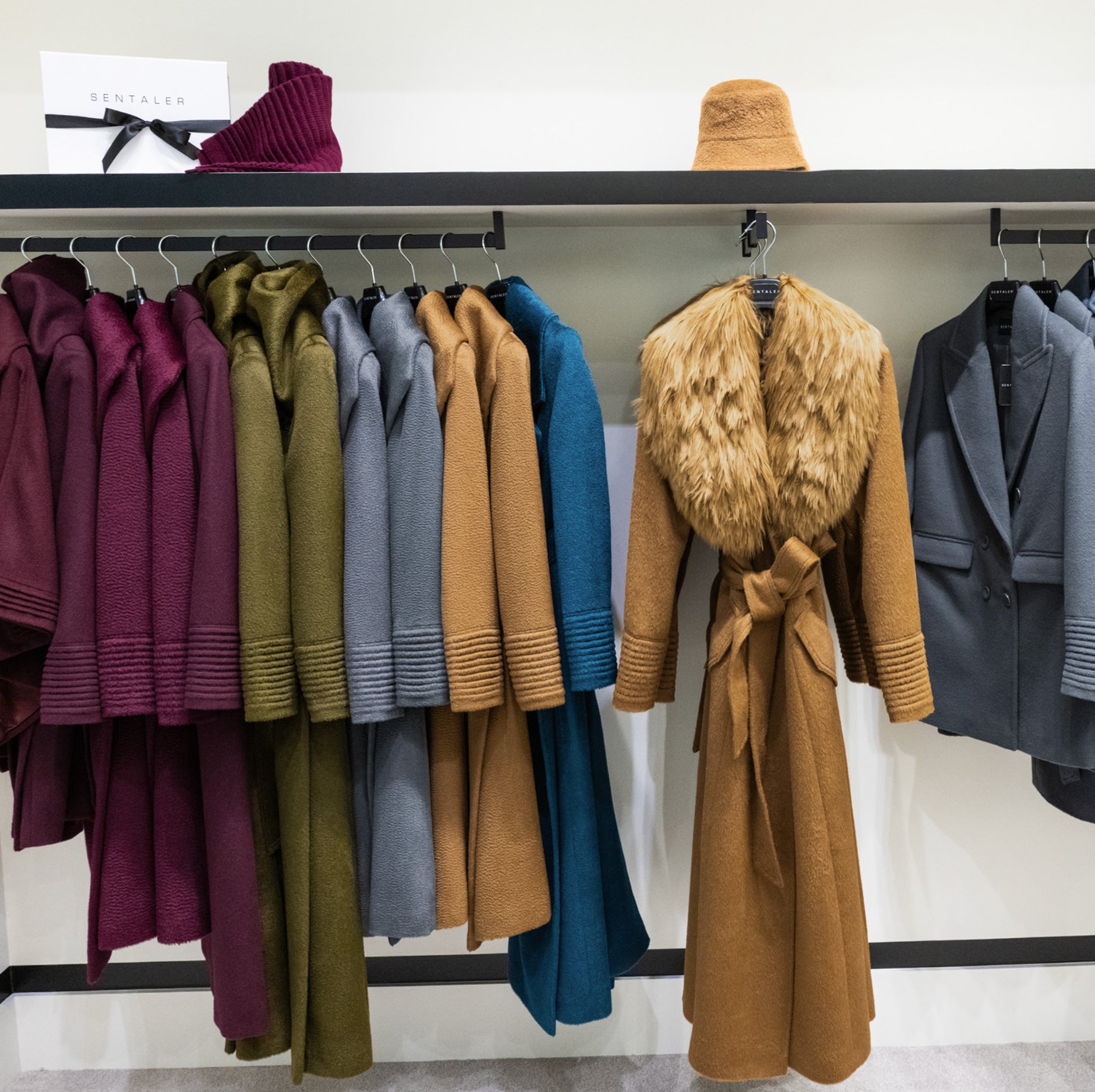 Display rack inside the SENTALER store featuring luxury alpaca wool coats in various colors including cherry lacquer, olive, dark camel, graphite grey, and emerald green, with a caramel brown coat with fur trim centered under a matching hat.