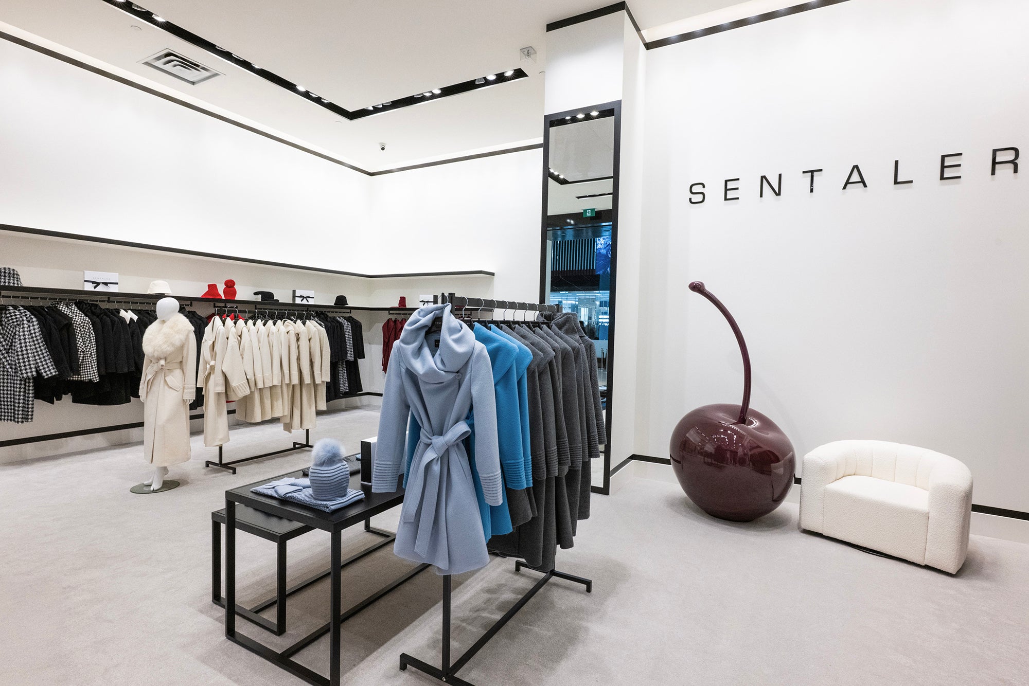 Interior of the SENTALER store showcasing neatly arranged racks of elegant wool coats in ivory, glacial blue, steel blue, graphite grey, black & black & white houndstooth, with a large glossy red burgundy cherry lacquer sculpture and a white armchair adding a modern design element.