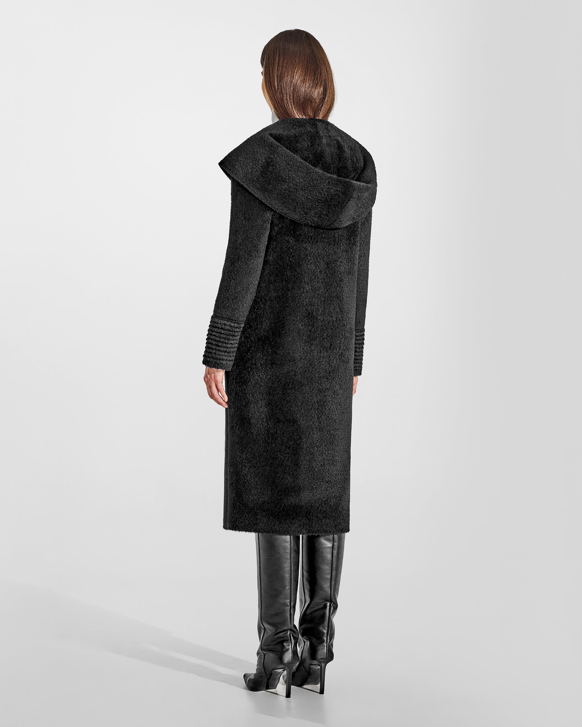 Back view of model wearing a Sentaler long black coat made of bouclé alpaca wool, with a hooded collar, worn buttoned.
