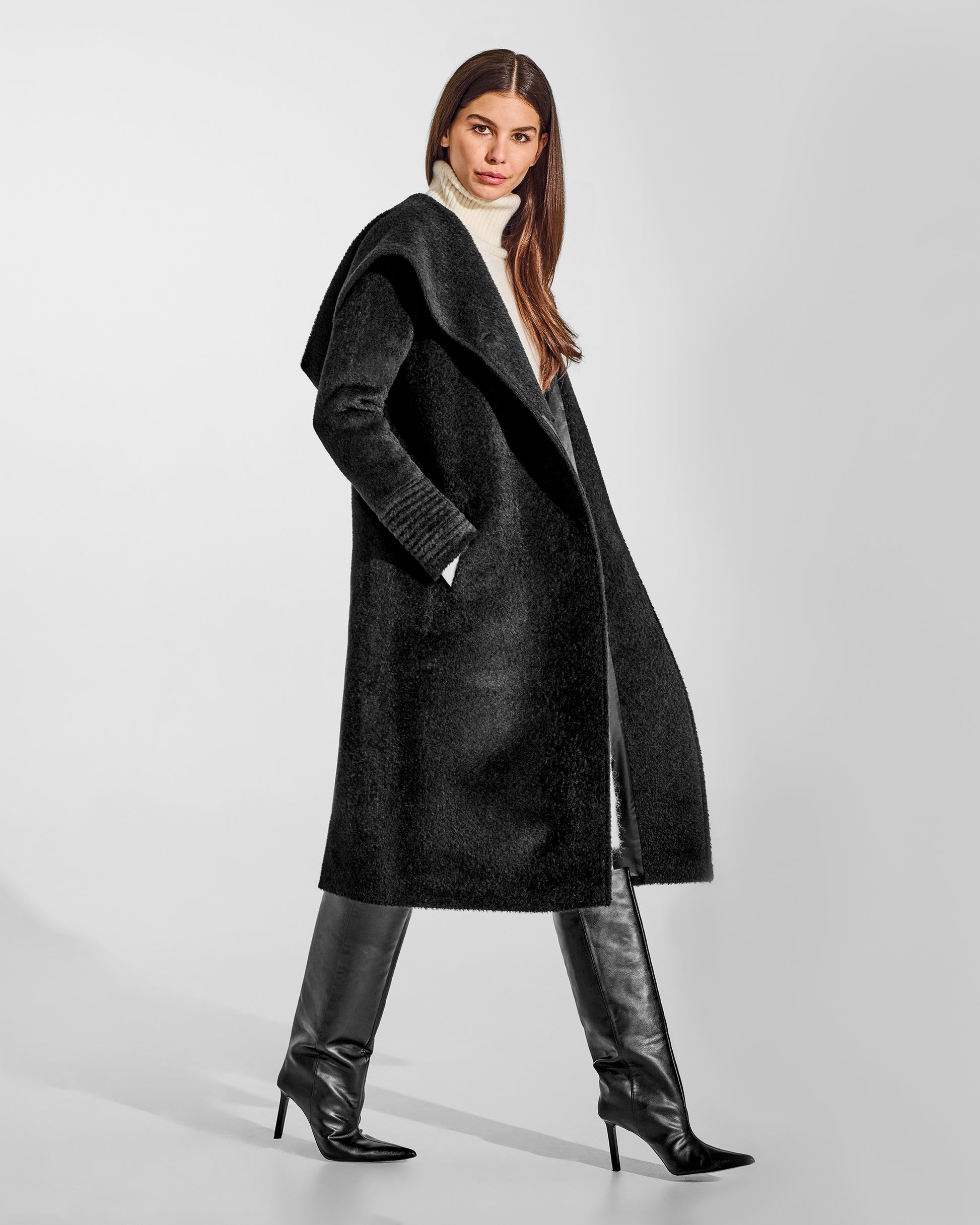 Side view of model wearing a Sentaler long black coat made of bouclé alpaca wool, with a hooded collar, worn open.